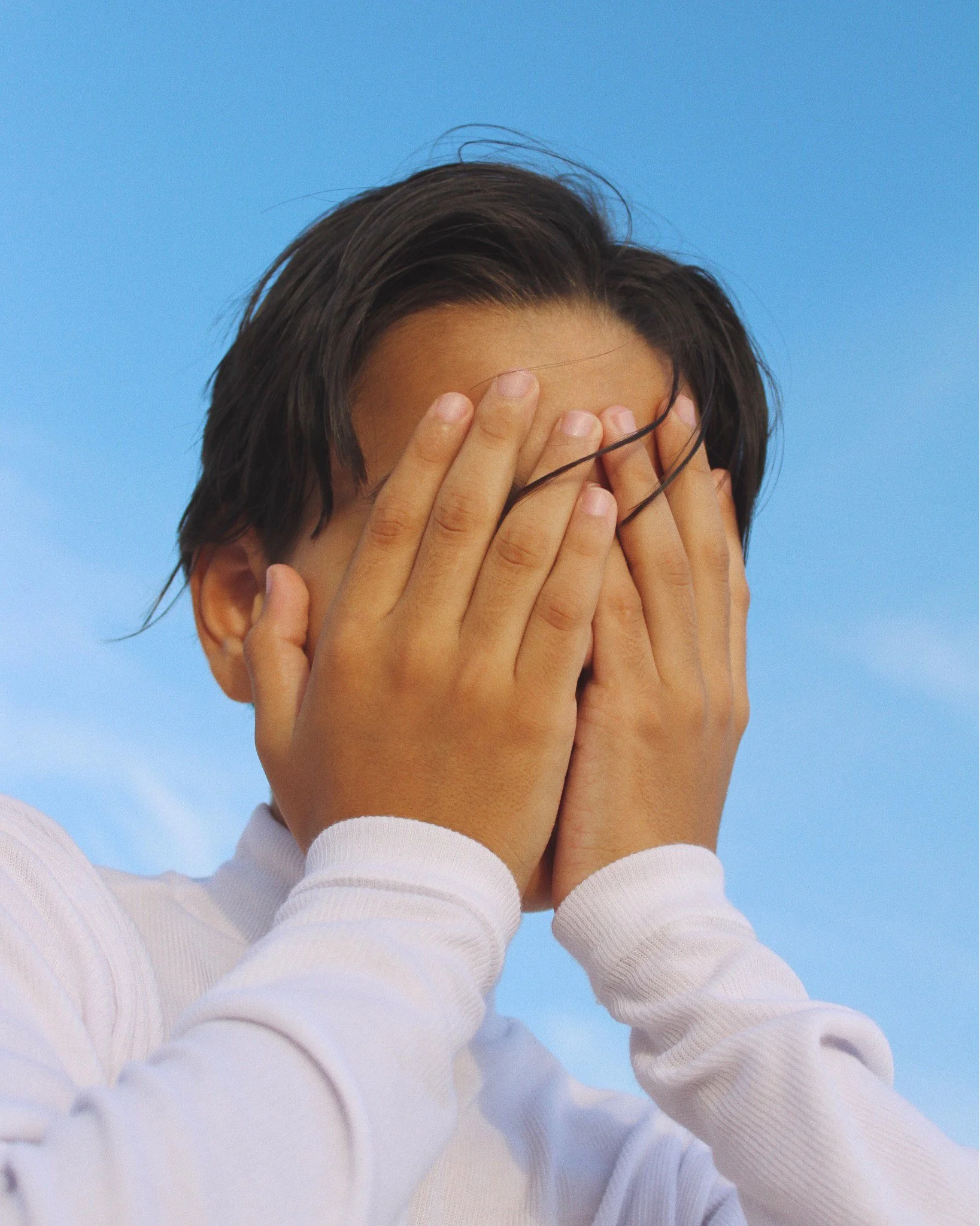 Person with dark hair covering their face with their hands, wearing a white long-sleeve shirt against a blue sky background.