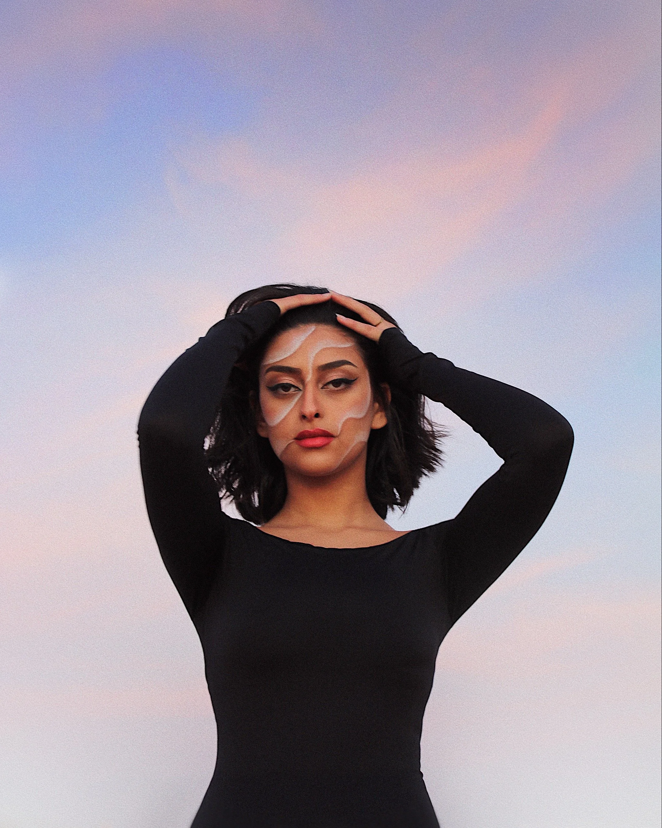 A woman with natural makeup and black hair, wearing a black long-sleeve dress, standing outdoors against a pastel sky, with her hands on her head, looking confident.