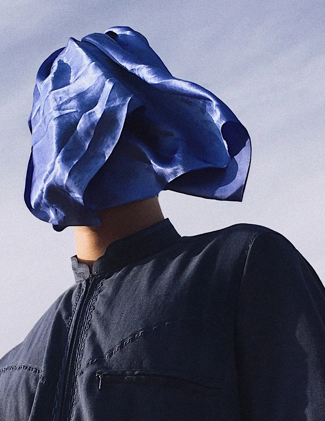 Person with a blue fabric covering their head, wearing a dark jacket, against a cloudy sky.
