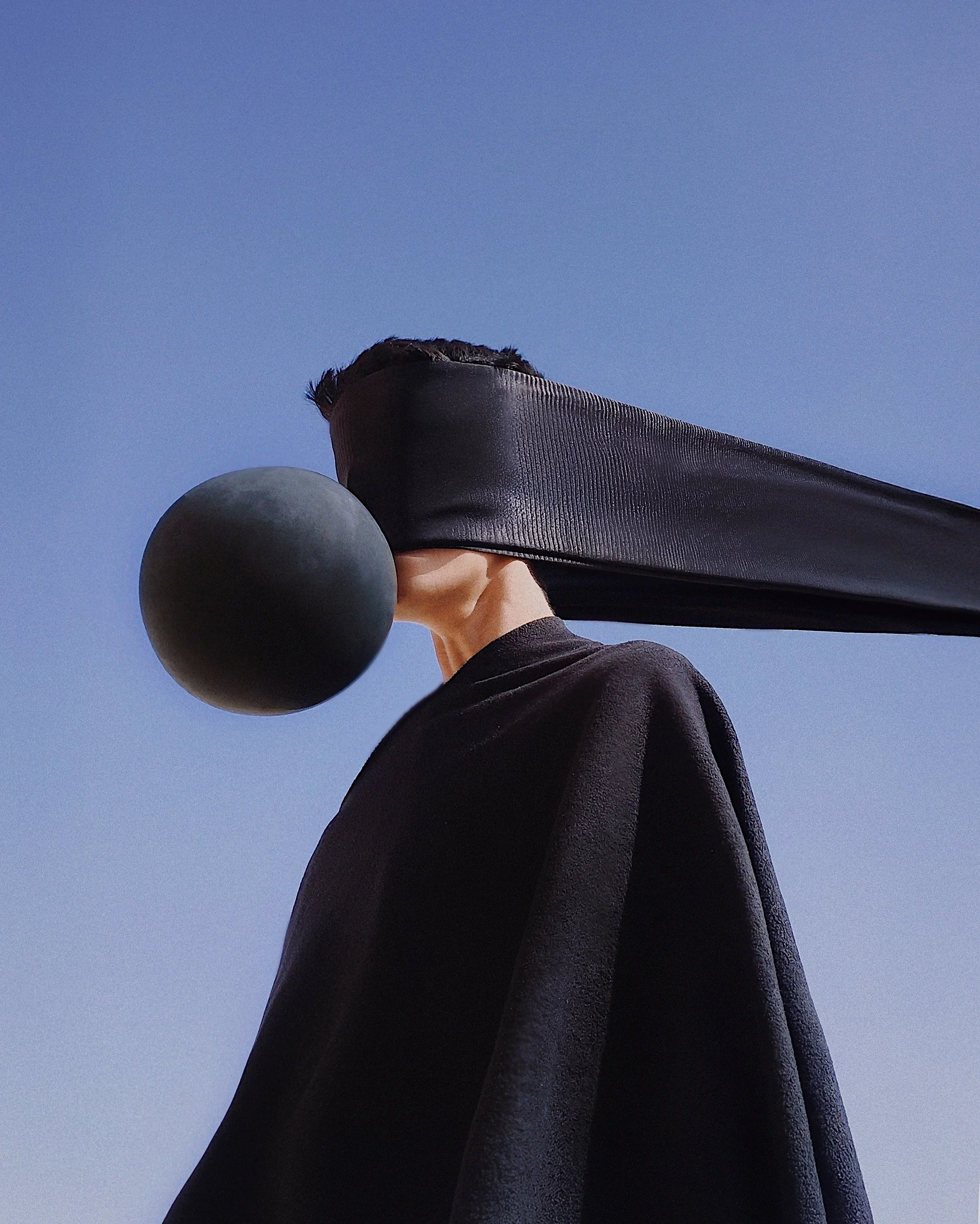 A person wearing black clothing and a blindfold carrying a black ball on their shoulder against a clear blue sky.