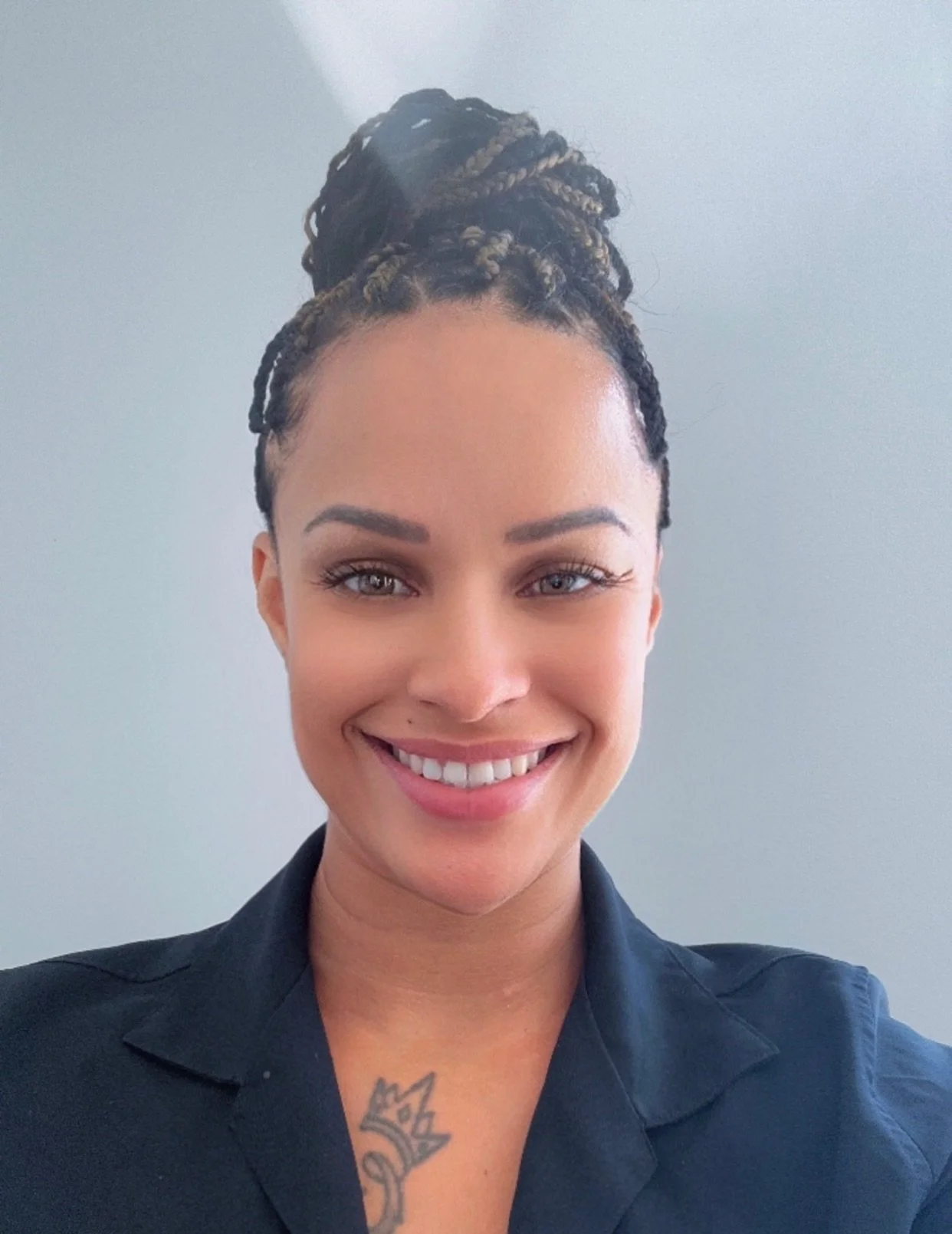 A woman with braided hair styled in an updo, wearing a black blazer, smiling in front of a plain background.