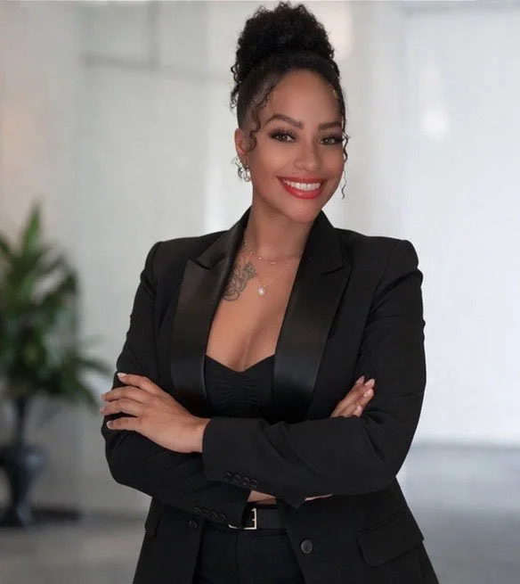 A confident woman with dark curly hair styled in an updo, wearing a black blazer over a black top, smiling and crossing her arms indoors with a blurred background.