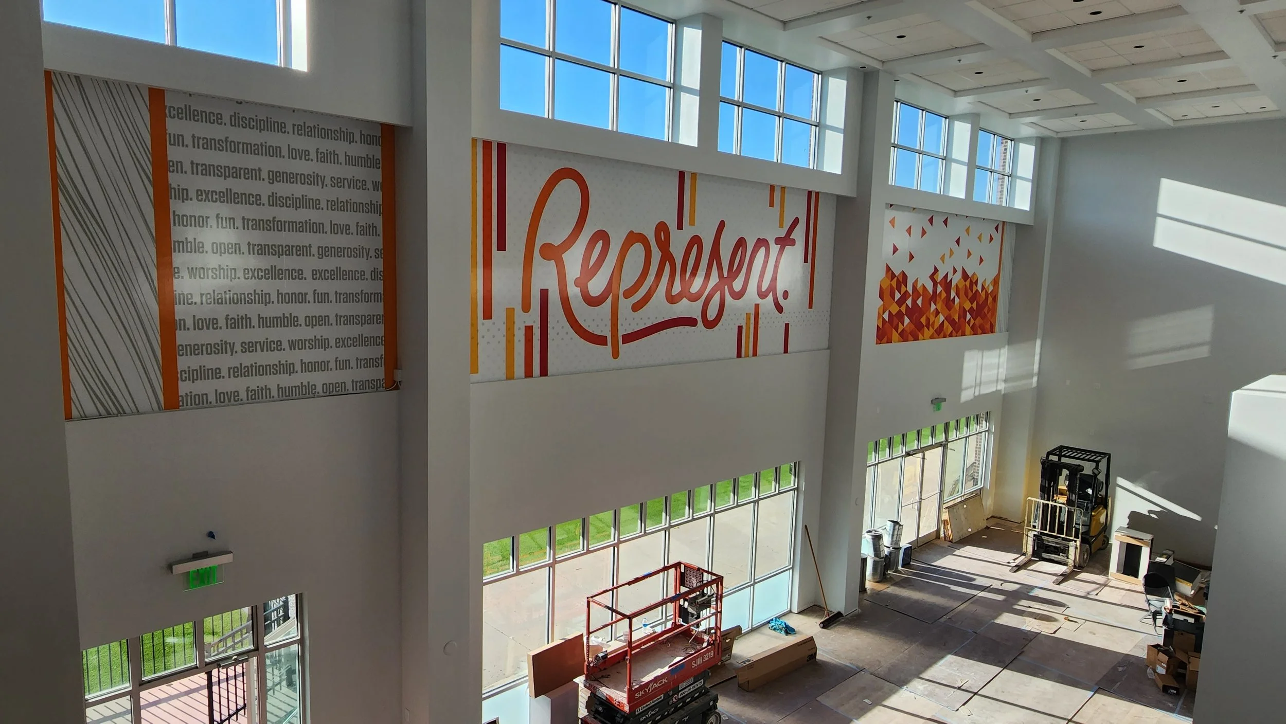 Interior of a large, bright room with high windows, featuring wall art with the word "Represent" and motivational text. Construction equipment like a lift and boxes are on the floor, suggesting ongoing work or setup.
