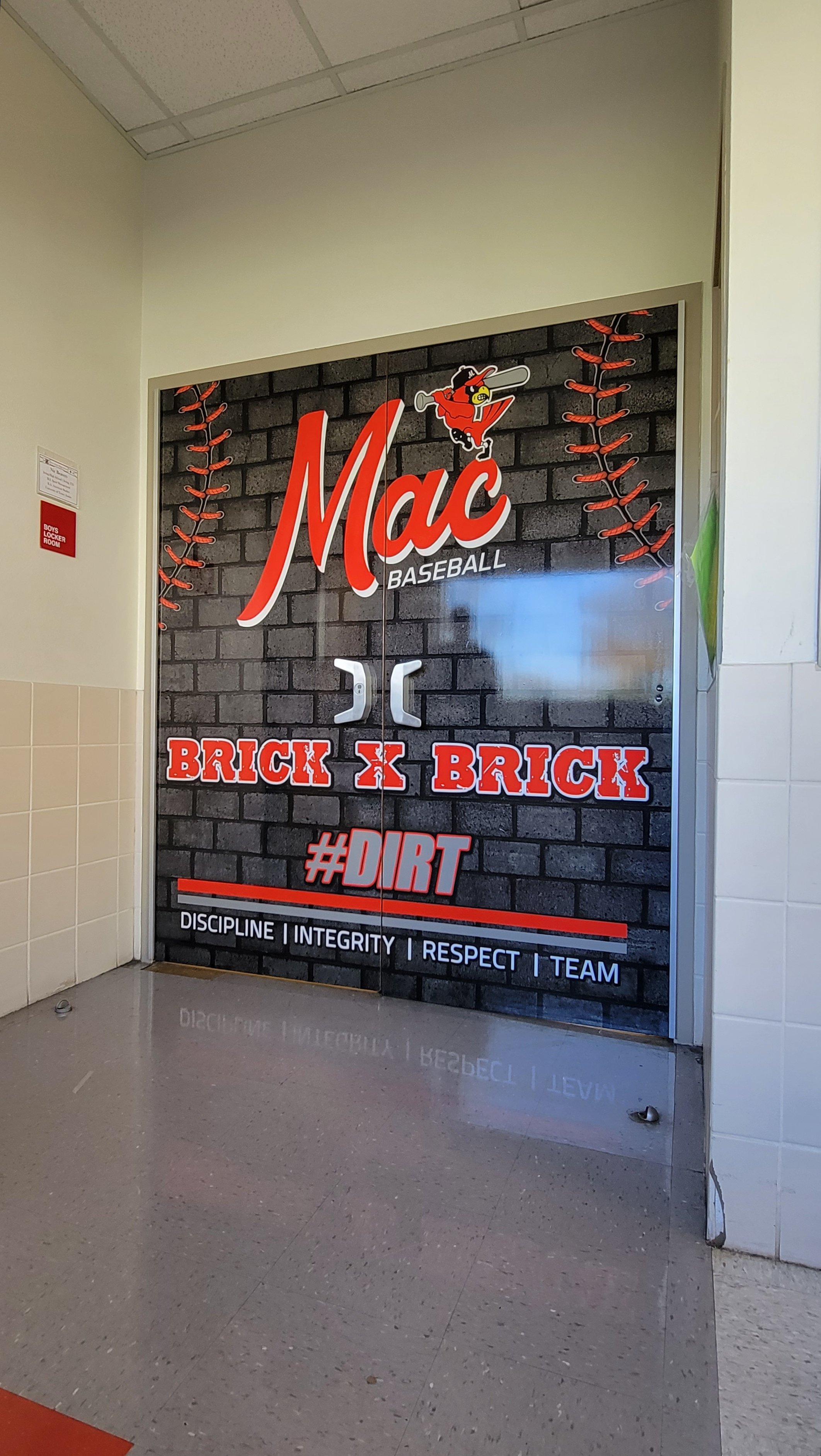Entrance to a baseball team locker room with 'Mac Baseball' signage, featuring graphics of a baseball and bat, labeled 'Brick x Brick,' '#DIRT,' and values like 'Discipline,' 'Integrity,' 'Respect,' 'Team.'