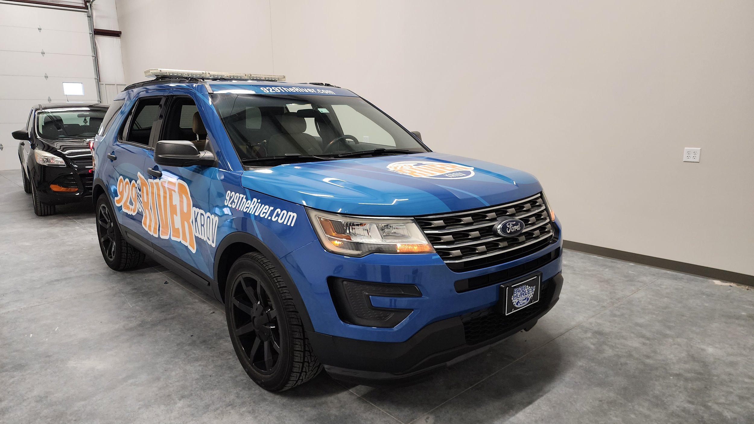 A blue Ford SUV with "92.9 The River" branding parked indoors, next to a black car.