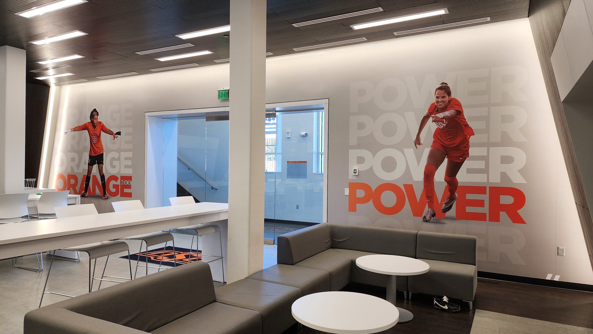 Modern indoor sports lounge with large wall graphics of soccer players, featuring the words 'ORANGE' and 'POWER', and furniture like tables, chairs, and sofas.