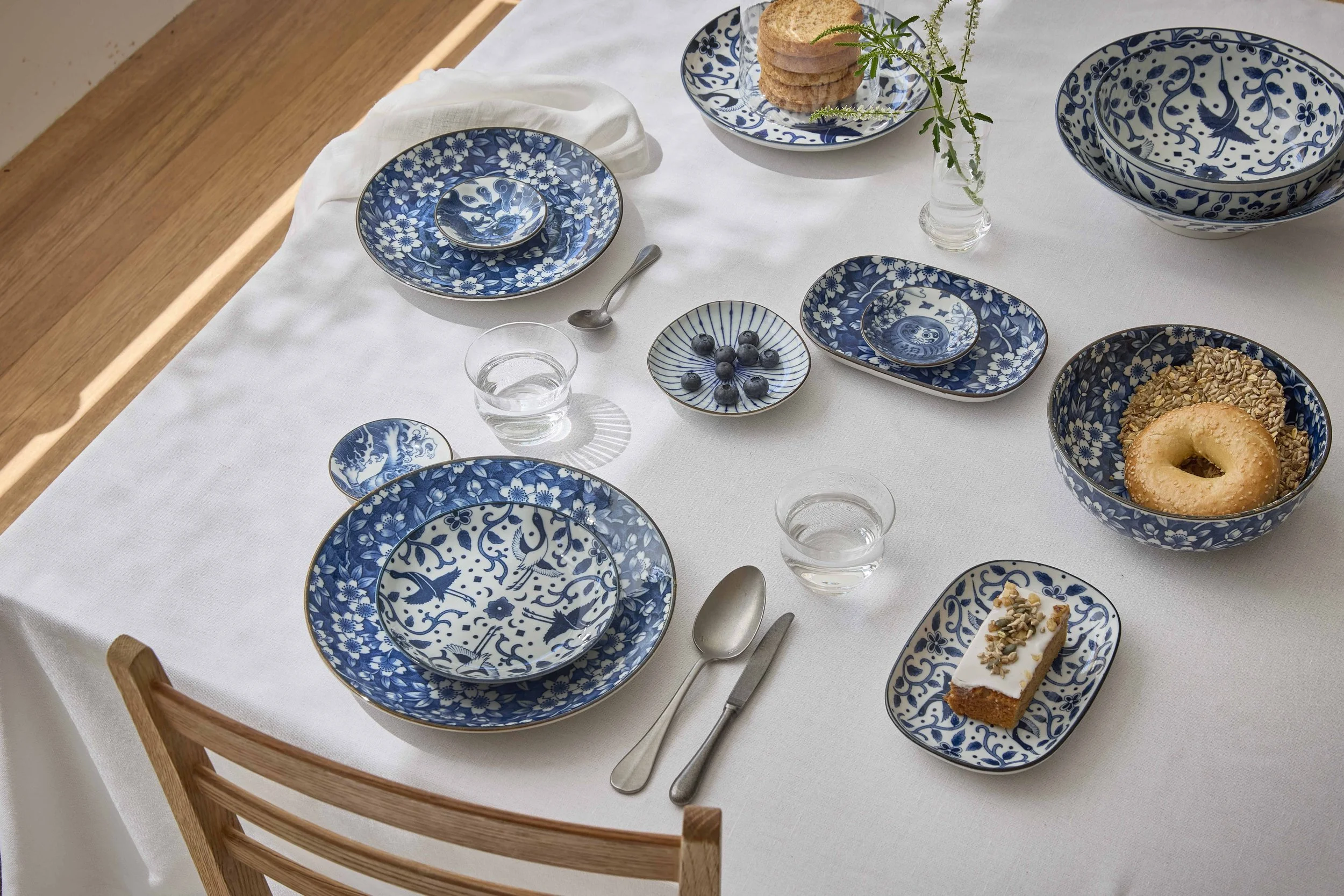 Tokyo Design Studio blue and white porcelain tableware set on a breakfast table — plates, bowls and dishes with floral and crane motifs, styled with bread, bagels and fruit