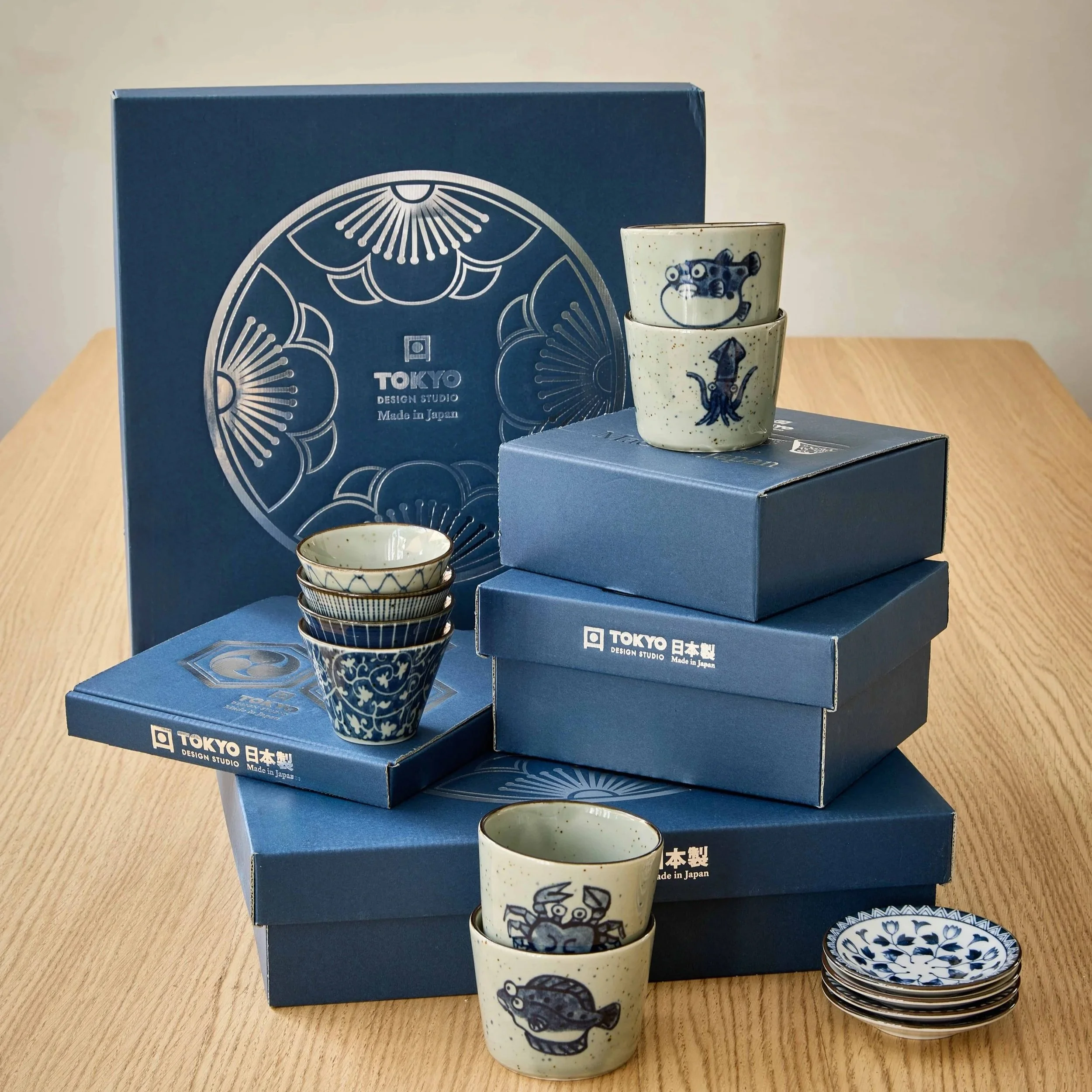 Tokyo Design Studio packaging with Japanese ceramic tableware — glazed cups featuring crab, fish and squid illustrations, and blue-and-white patterned bowls, displayed with branded navy gift boxes