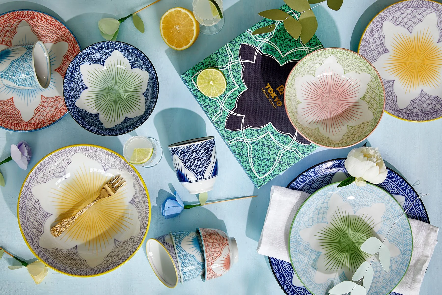 Tokyo Design Studio colourful floral patterned tableware — bowls and plates in blue, green, coral and yellow, styled with citrus, flowers and branded packaging on a light blue background