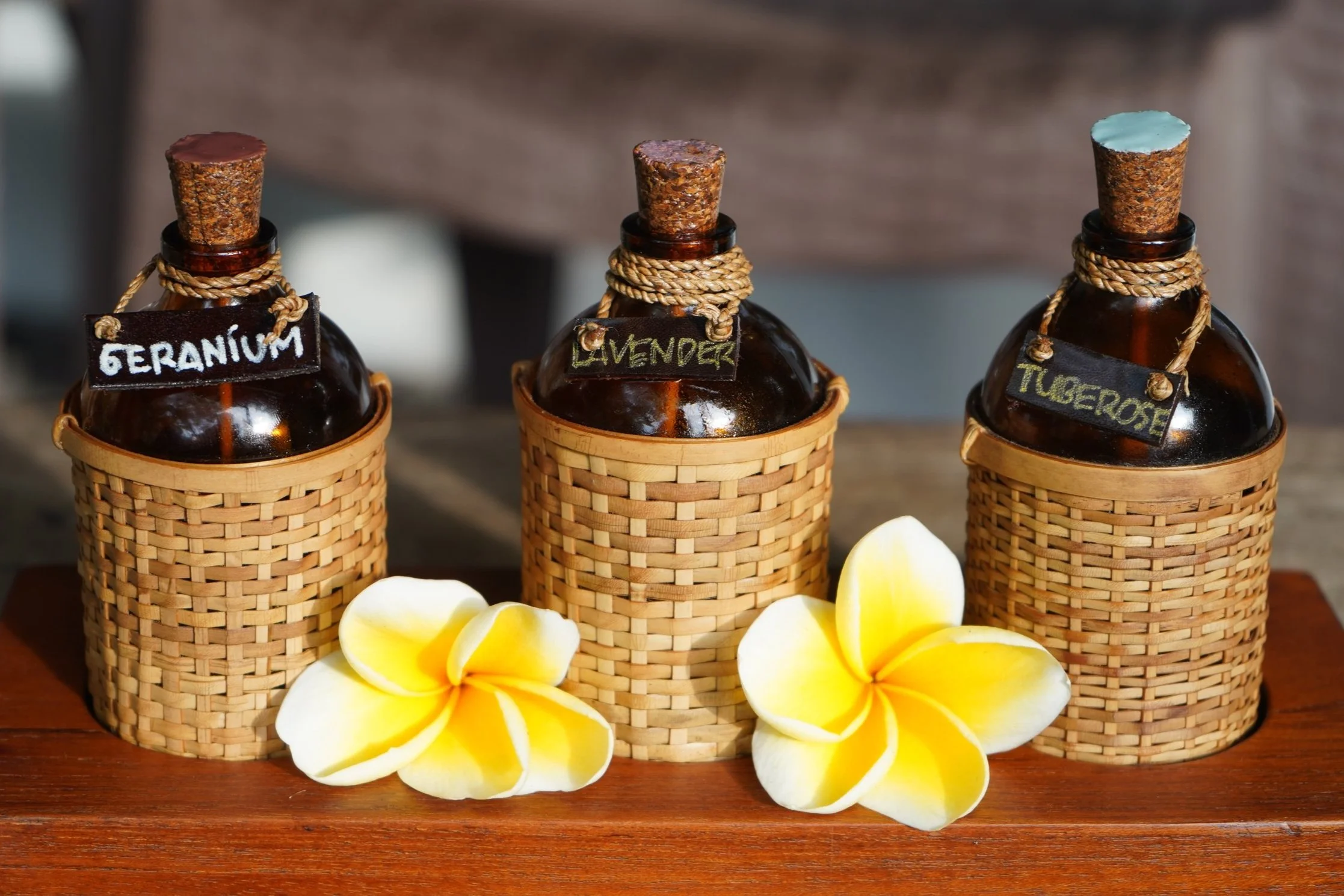 Three glass bottles with cork stoppers labeled geranium, lavender, and tangerine, each nestled in woven basket holders, with yellow and white plumeria flowers in front on wooden surface.