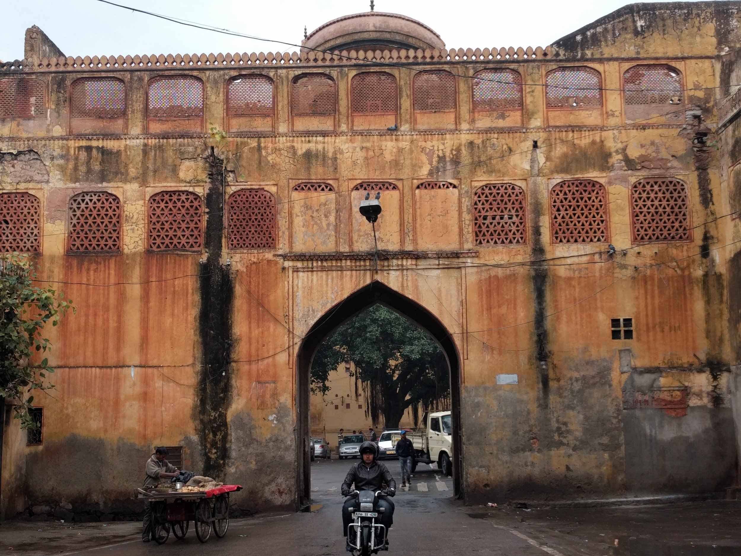 Ancient yellow and orange fortress with arched gate, lattice windows, and people riding a motorcycle and selling goods on a cart outside.