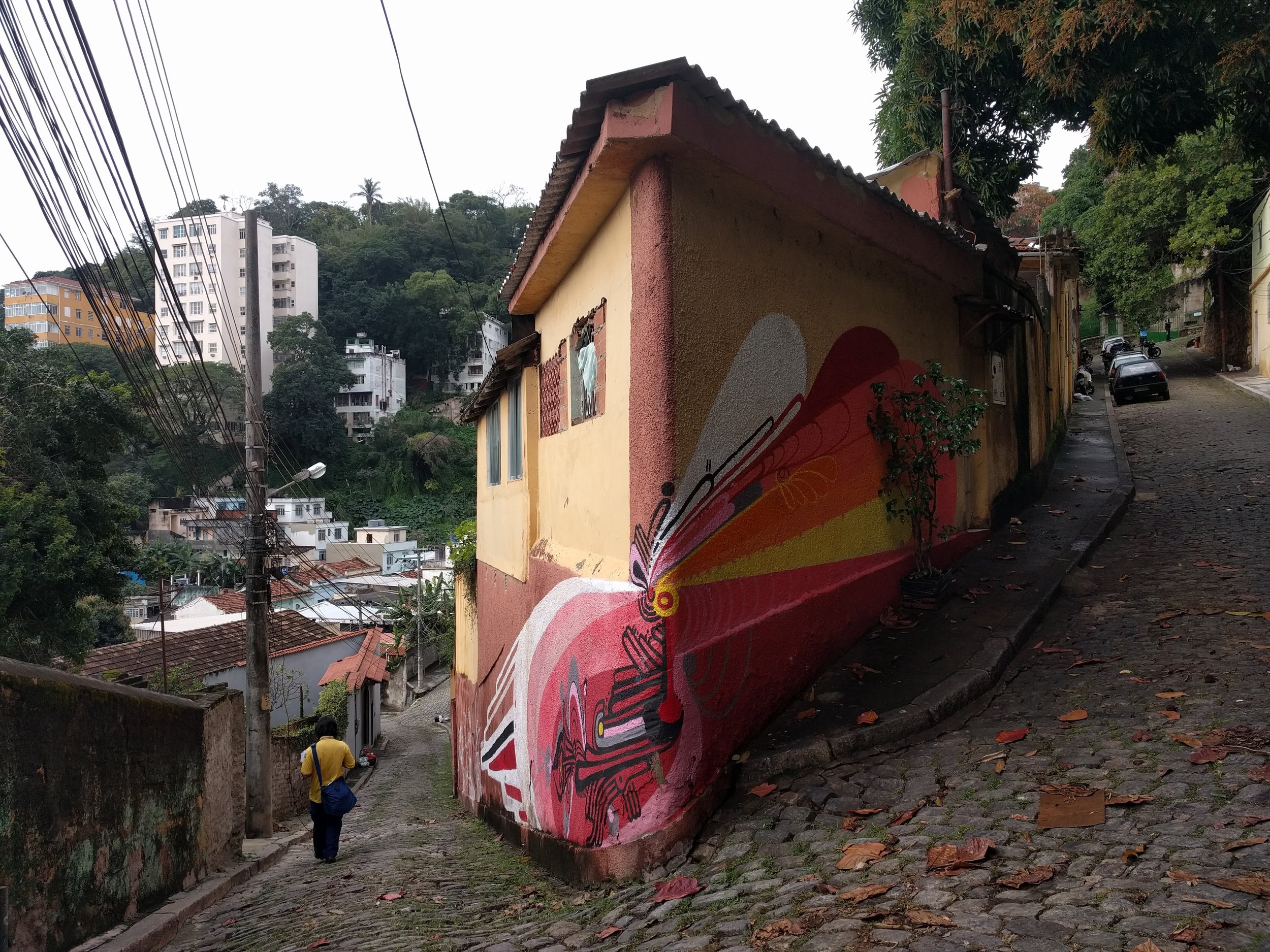 A cobblestone street on a hillside in a neighborhood, with a yellow house featuring colorful street art on its wall, and taller modern buildings in the background. A person with a yellow shirt and blue bag walks down the street.