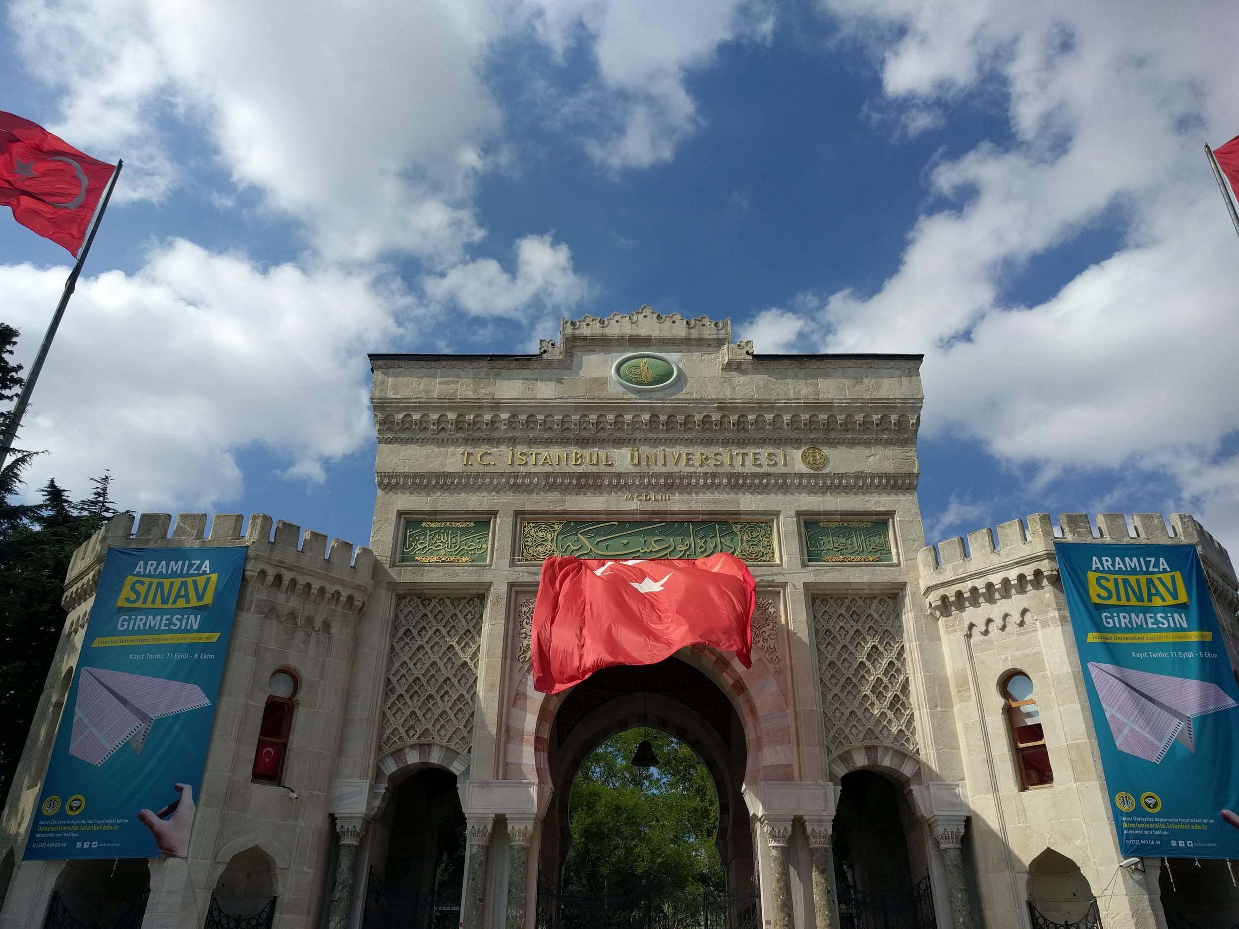 The entrance gate of T.C. Istanbul University, decorated with Turkish flags and banners, with a blue sky and clouds in the background.