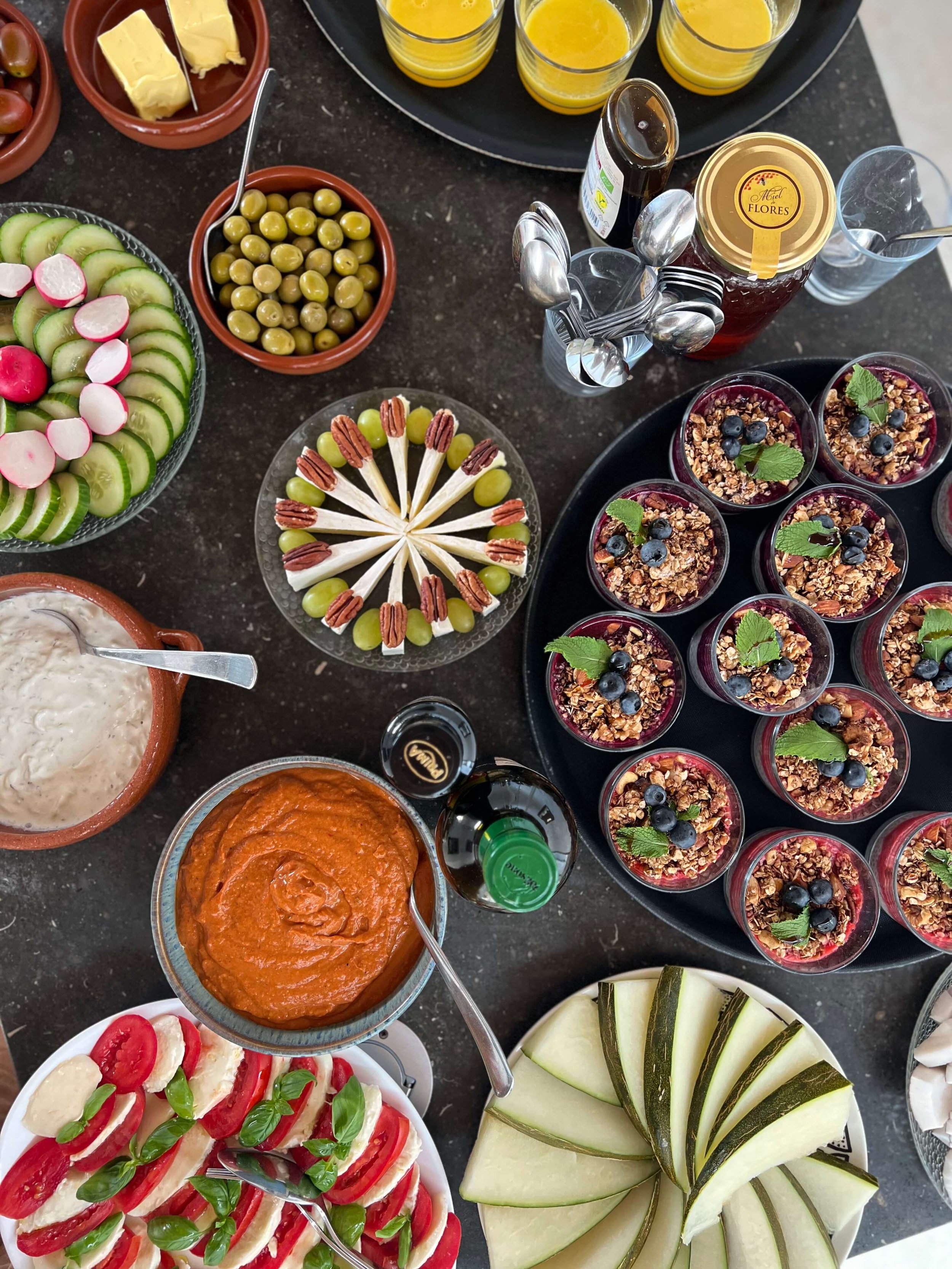 Verschiedene Frühstücks- und Dessertgerichte auf einem Buffet, einschließlich Gurken- und Radieschensalat, grünen Oliven, Mini-Sandwiches mit Tomaten, Melonenscheiben, Joghurt mit Granola und Beeren, Chutney, Curry, Müsli mit Beeren und Minzblättern,