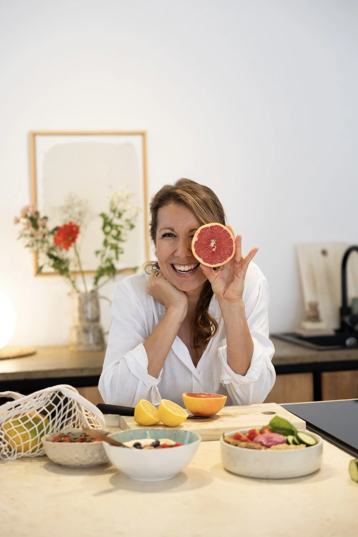 Frau sitzt am Küchentisch, hält eine halbe Grapefruit vor ihr Gesicht und lacht, umgeben von Obst und Salat in Schüsseln.