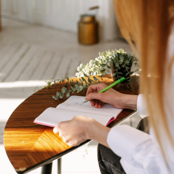 Person schreibt in ein Notizbuch auf einem Holztablett mit grünem Stift, neben einer Pflanze auf der Tisch.