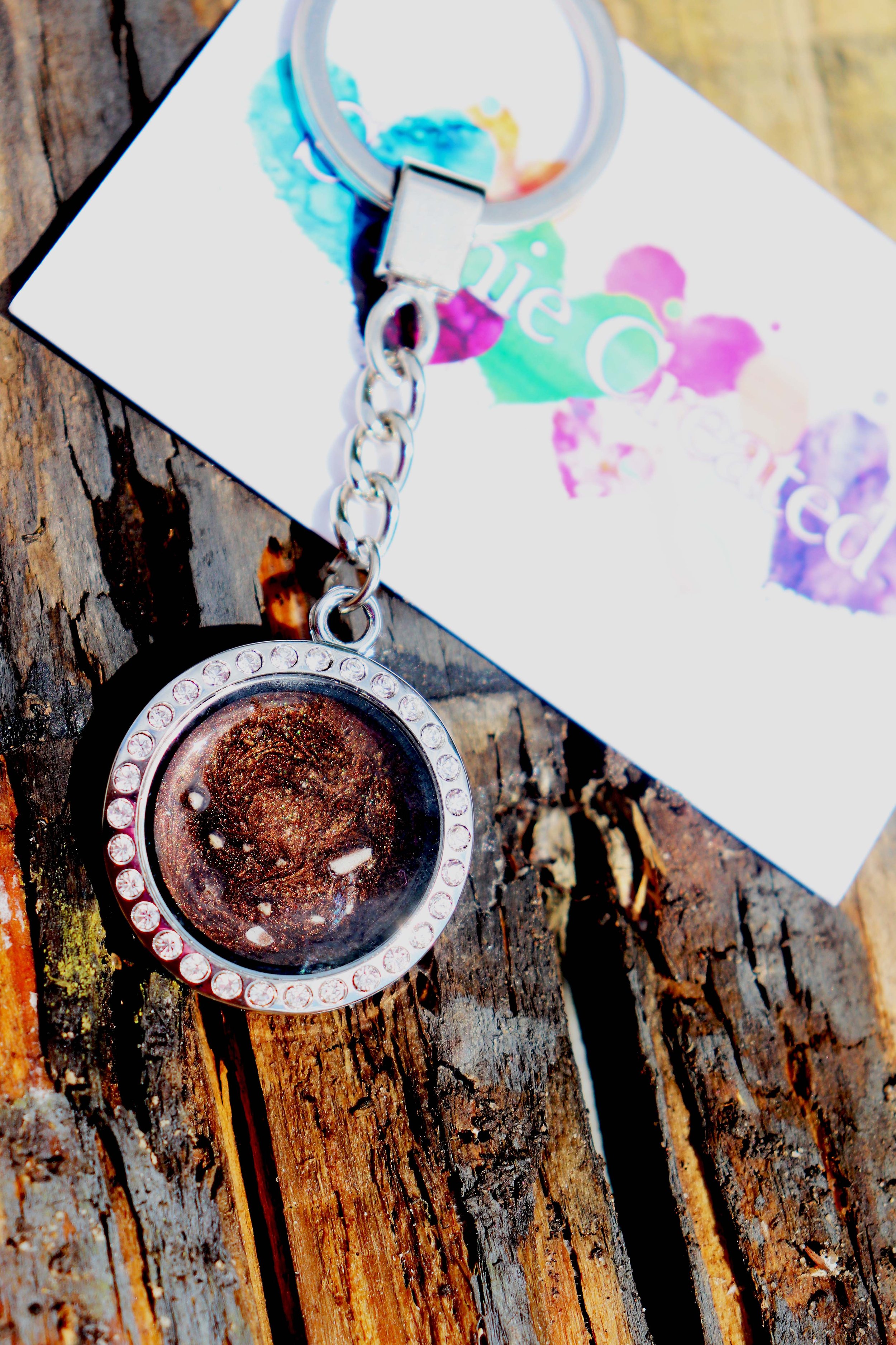 A keychain with a circular glass pendant containing glitter and small white and pink objects, resting on a wooden surface with a colorful watercolor-painted card underneath.