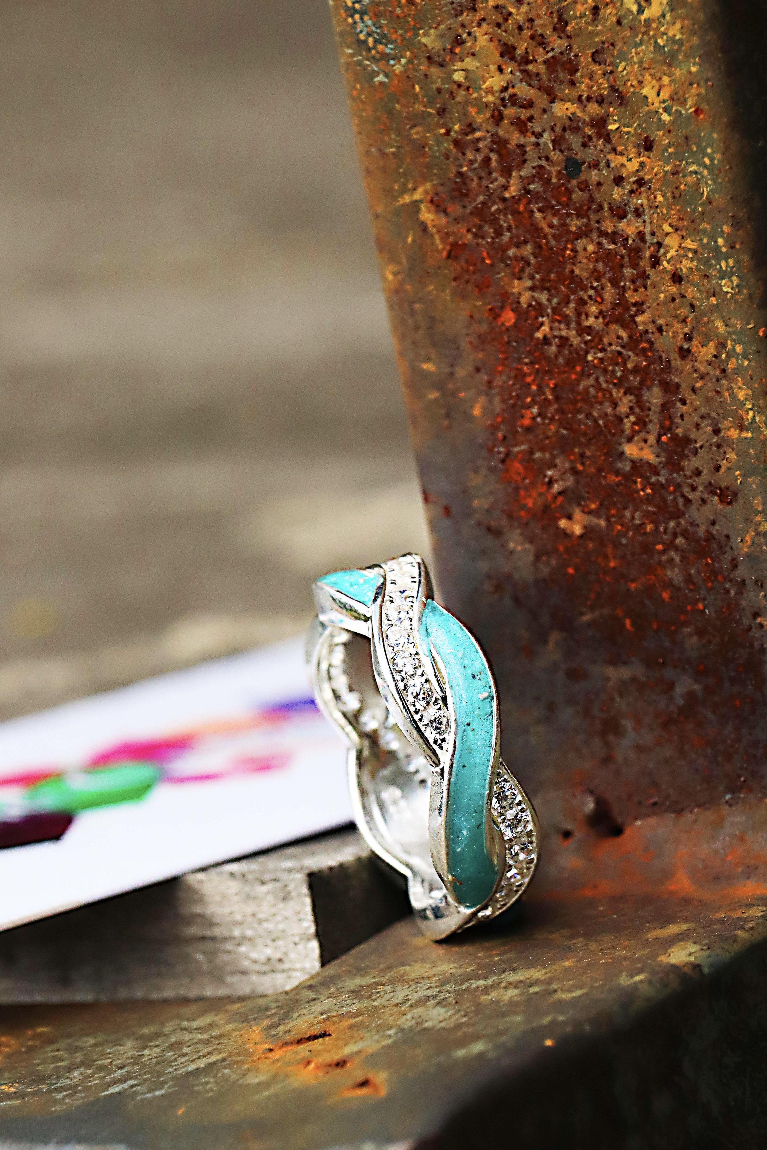 A silver ring with blue enamel and embedded diamonds leans against a rusted metal surface, with a blurred background.