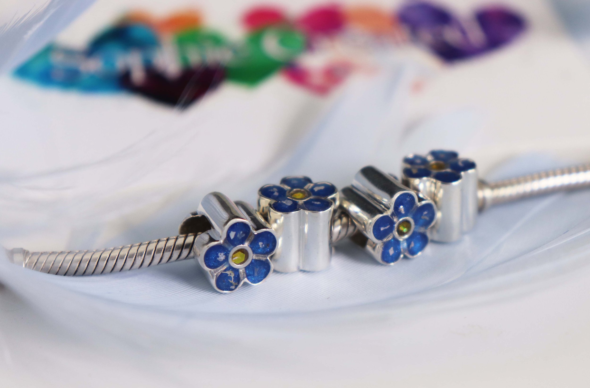 Silver bracelet with blue flower charms on a white fabric surface, with a colorful blurred packaging in the background.