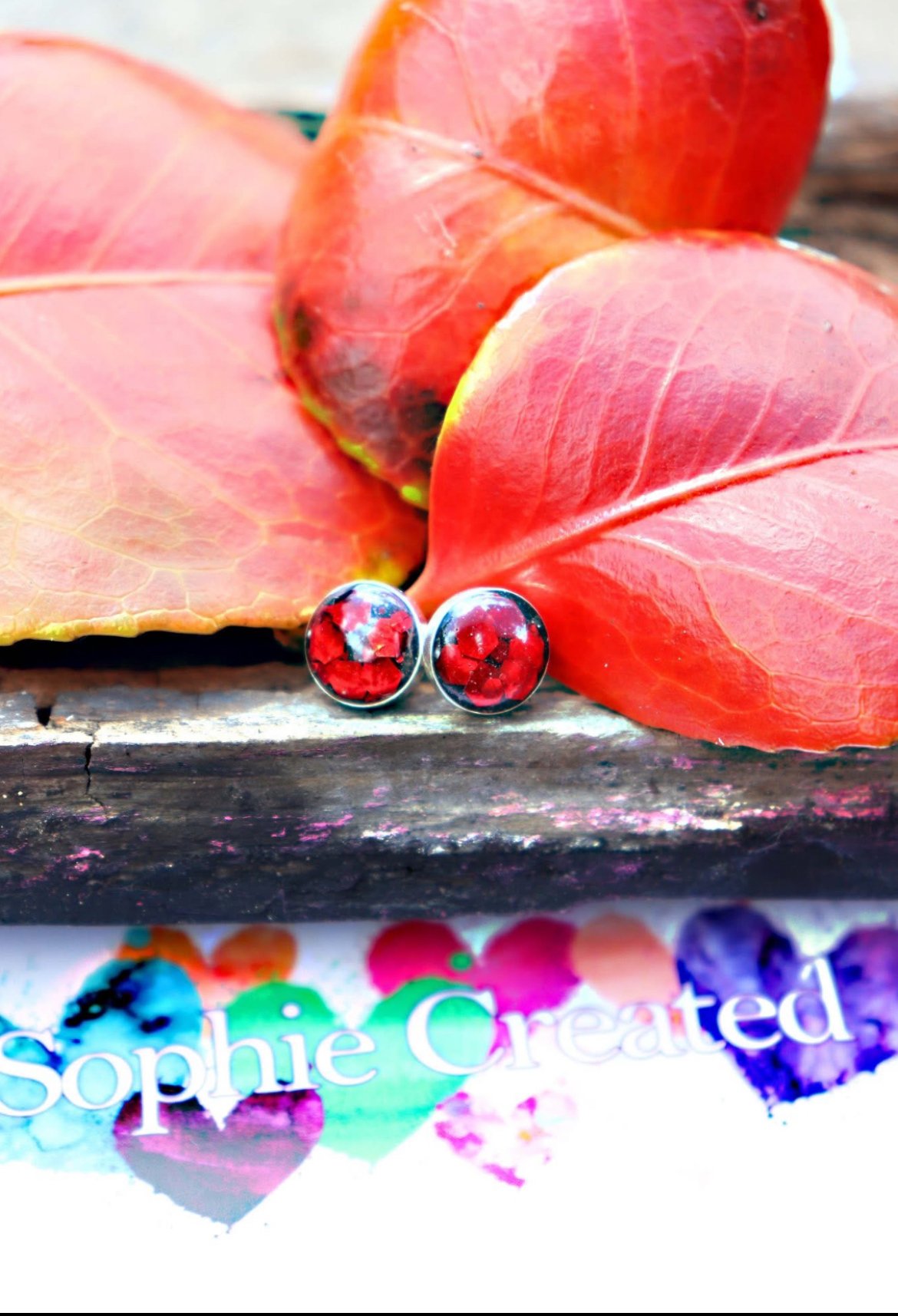 A pair of glass earrings with red and black patterns resting on red autumn leaves, placed on a wooden surface. Part of a book cover with the text 'Sophie Created' and colorful heart shapes is visible at the bottom.