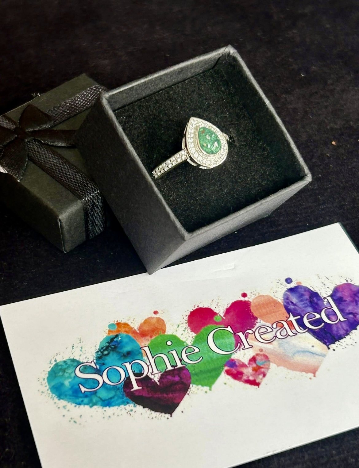 A jewelry ring with a green gemstone and diamonds in a black box, next to a colorful card with the name 'Sophie Created' written on it.