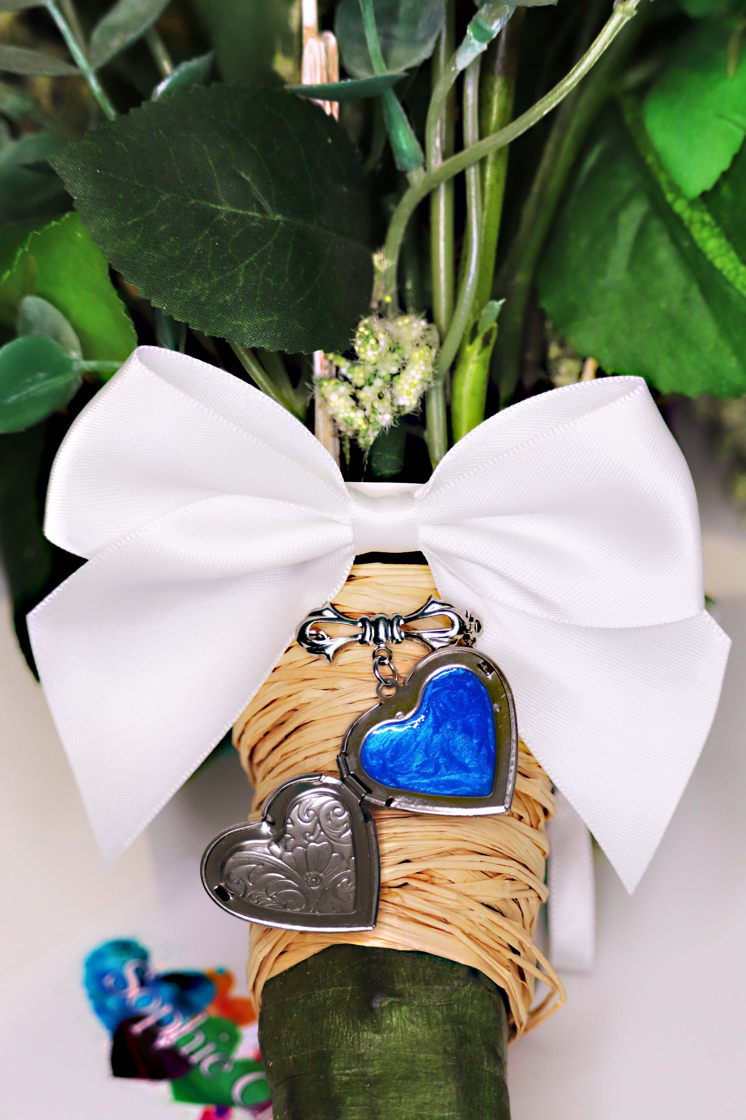 A decorated flower with a white bow and two heart-shaped charms, one blue and one silver, hanging from a green stem with leaves.