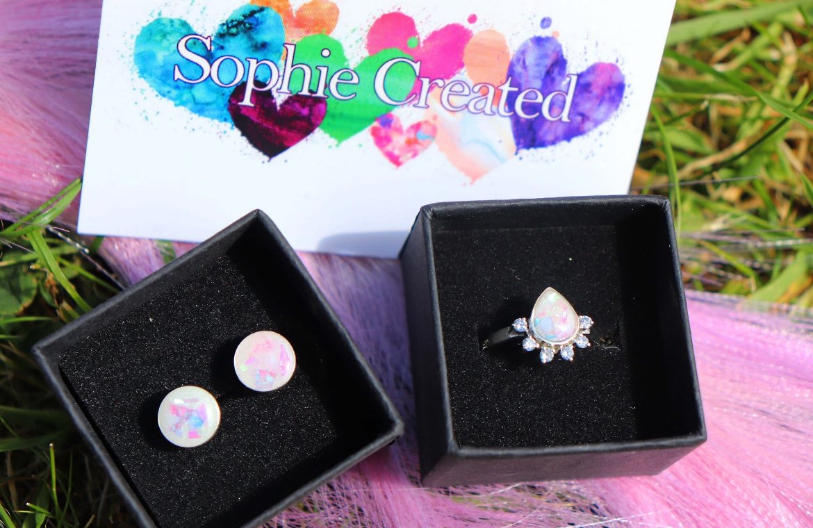 A pair of opal stud earrings and a opal and diamond ring in black jewelry boxes, placed on pink fabric with grass around. A sign with the text 'Sophie Created' and colorful heart designs is in the background.