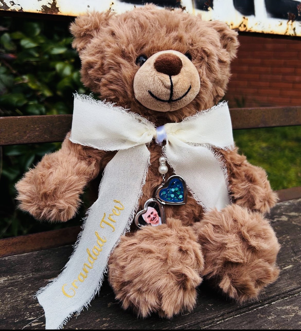 A brown teddy bear with a cream-colored nose and a white satin ribbon tied around its neck, sitting on a wooden bench. The bear has two necklaces with heart-shaped pendants, one with a photo of a smiling baby and the other with blue decorations. The