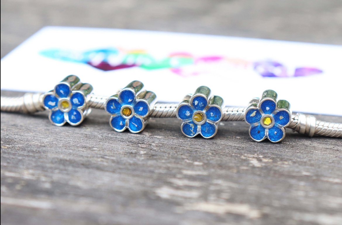 Four silver charm beads shaped like blue flowers with yellow centers, aligned on a silver bracelet, on a wooden surface with a blurred colorful card in the background.