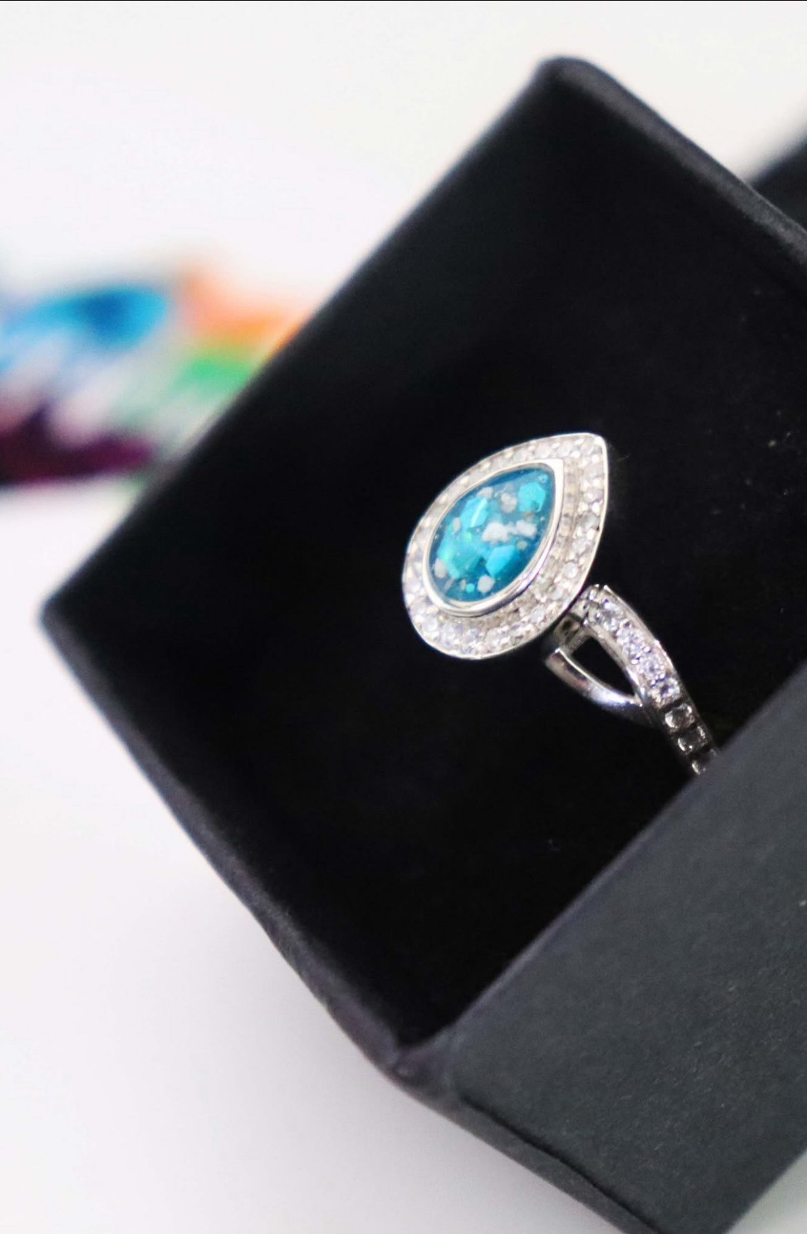 Silver ring with a large blue oval gemstone surrounded by small clear stones, placed inside a black jewelry box.