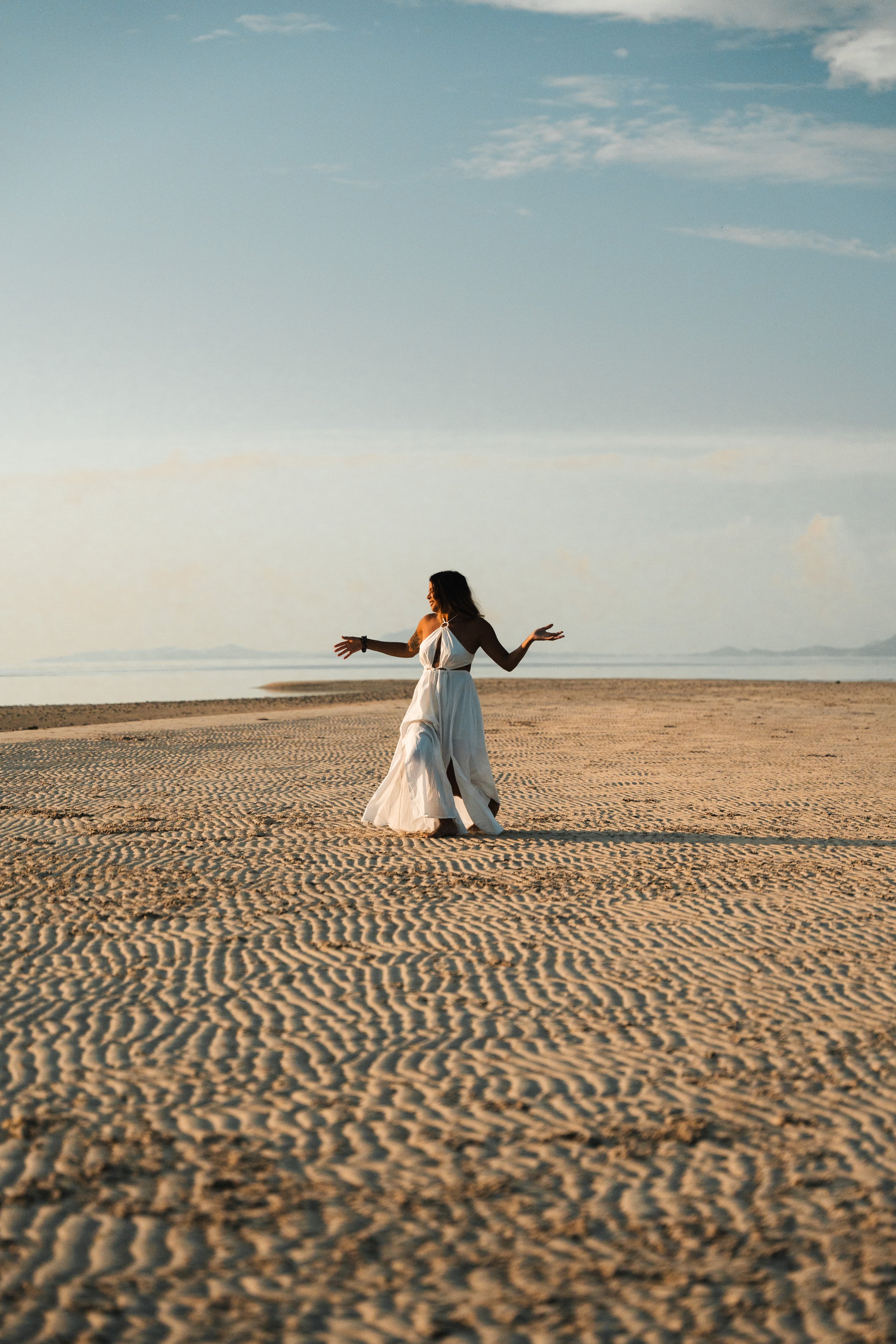 A woman in a white dress walking barefoot on a sandy beach during sunset.