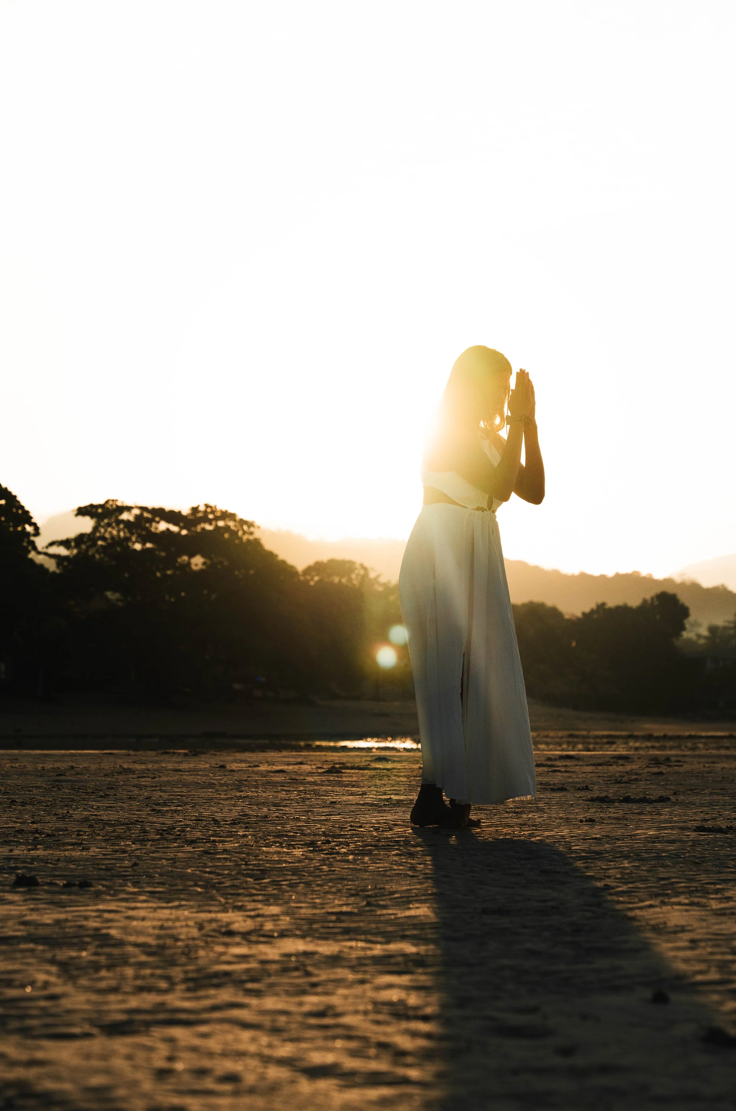 A woman standing outdoors during sunset, with her hands clasped together in prayer, wearing a white dress, and casting a shadow on the ground.