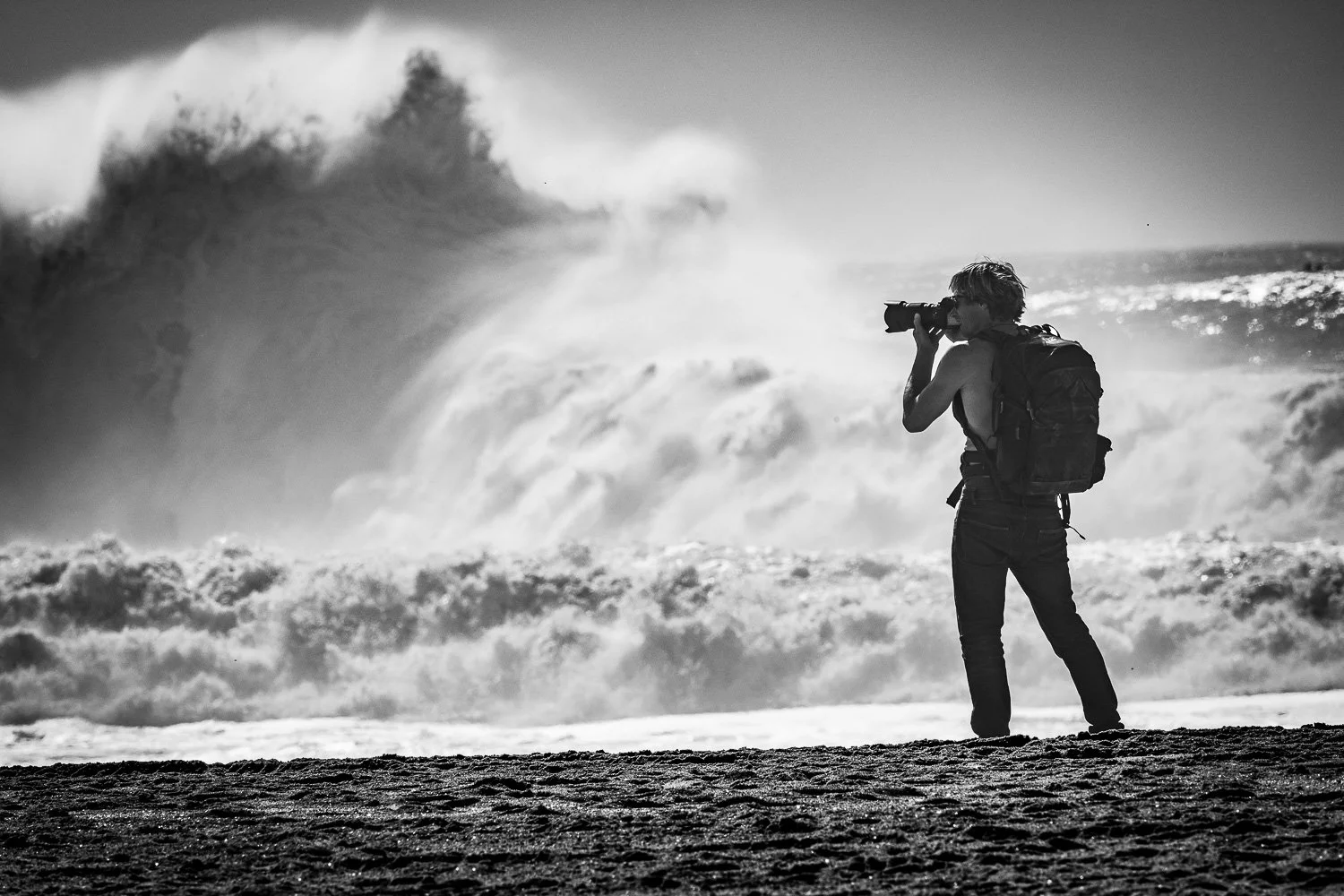 A person with a backpack standing on a sandy beach, taking a photo of the waves and ocean in black-and-white.