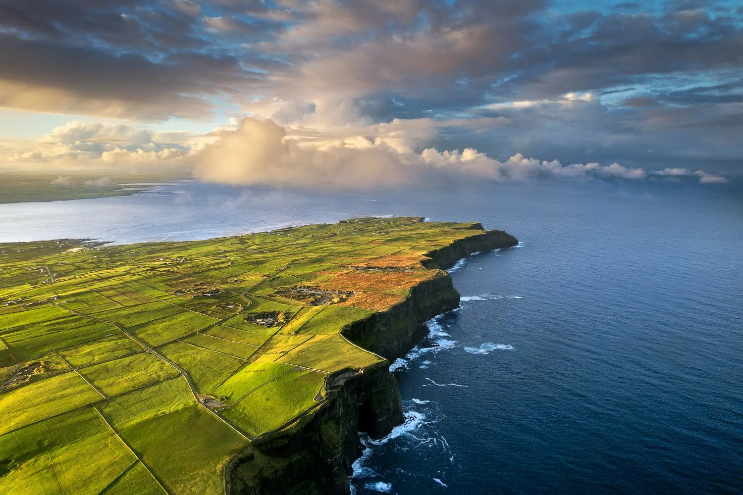 Aerial view of green fields of the Cliffs of Moher, a coastline with the ocean on the right and clouds in the sky.
