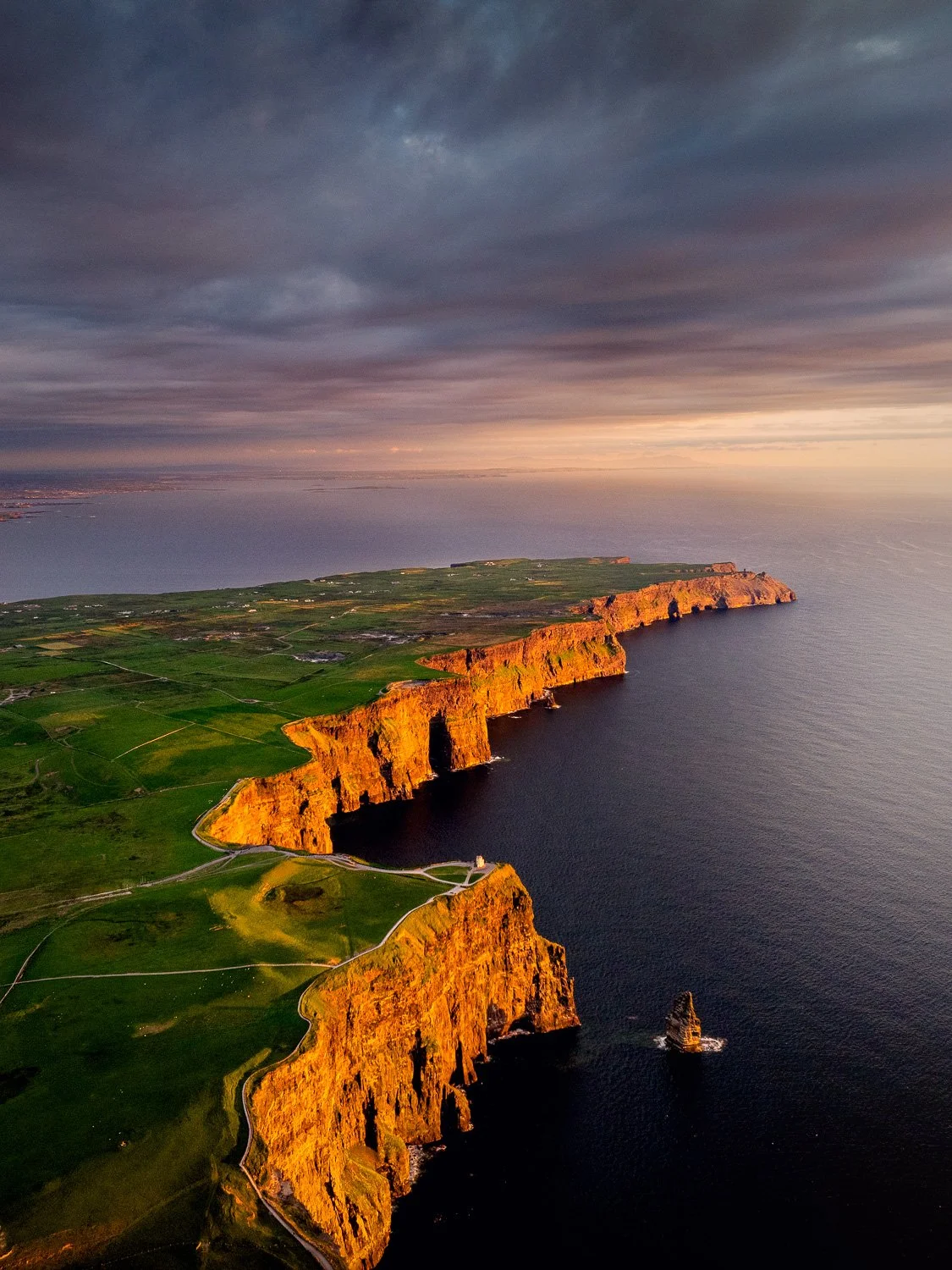 Aerial view of the Cliffs of Moher coastline with ocean and cloudy sky at sunset.