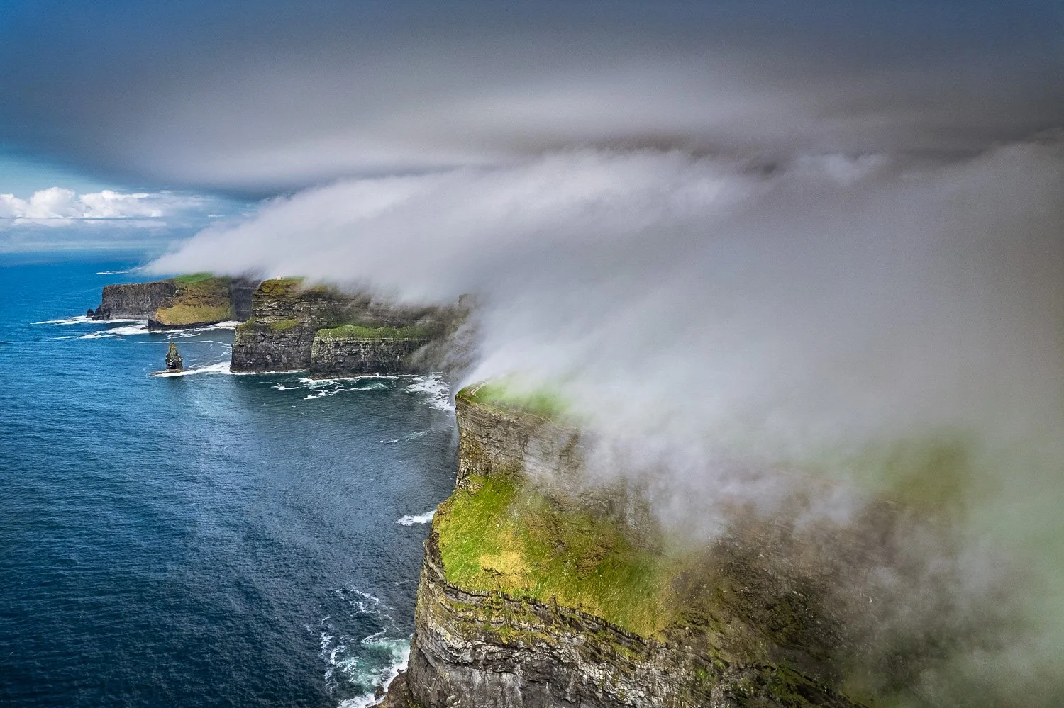 Cliffs of Moher coast with green grass patches, partially covered by fog and low clouds, with blue ocean water below.