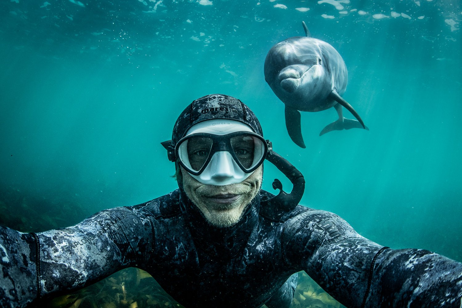 Underwater selfie of a free diver George Karbus framed by emerald light as a solitary bottlenose dolphin approaches from behind, creating a playful and spontaneous moment in the Atlantic.