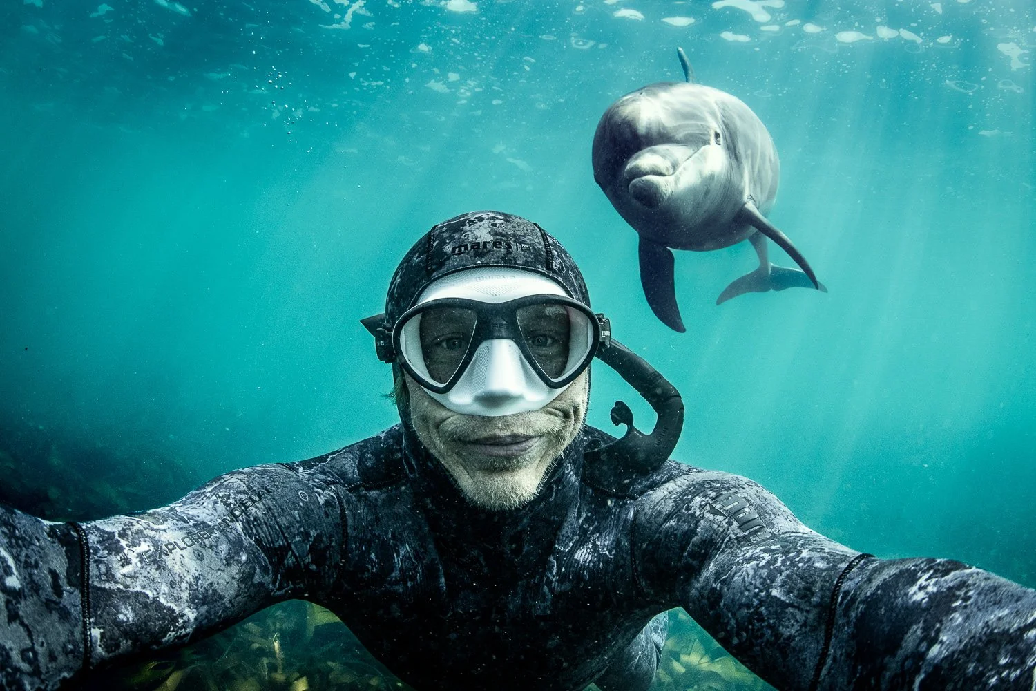 George Karbus takes a selfie underwater, with a dolphin swimming behind him in clear blue water.