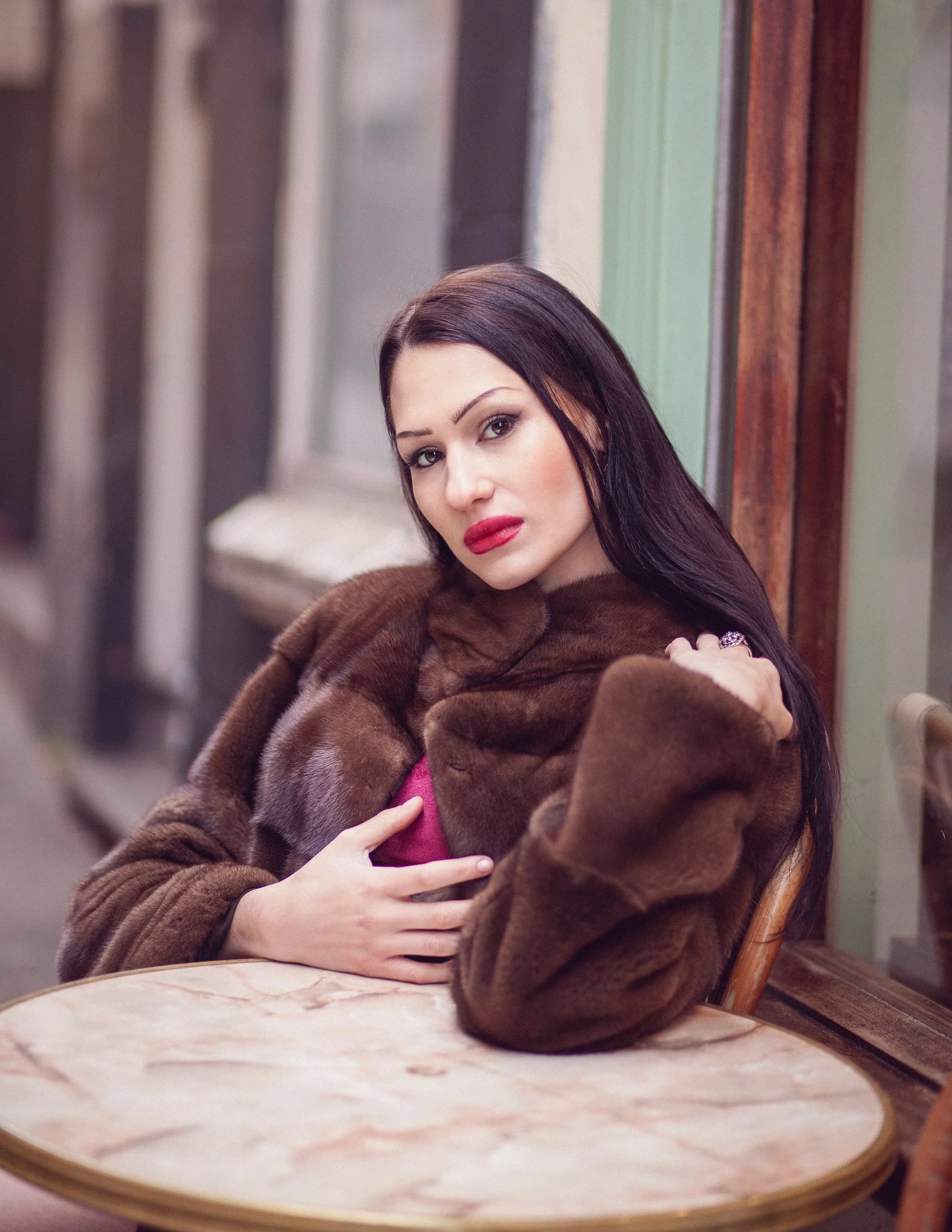 Luisa Young Editorial Photography in Nottingham of a woman with long dark hair, red lipstick, and a fur coat sitting at a table outside a café or restaurant, leaning on her hand and looking at the camera.