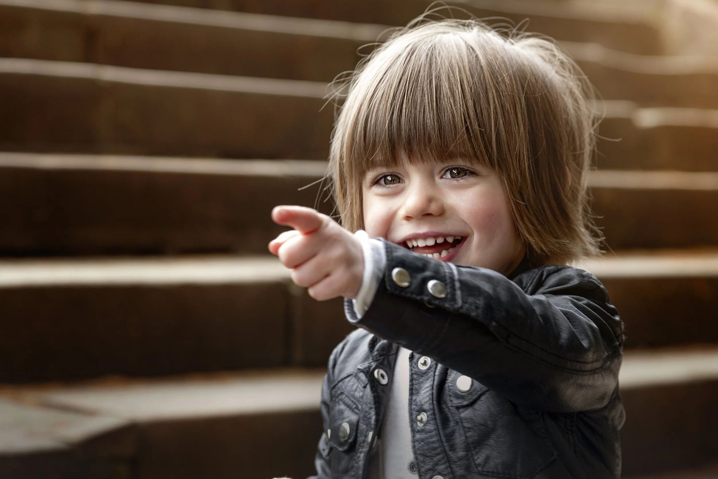 Young boy with blond long hair sitting on steps smiling and pointing his arm and finger towards the camera