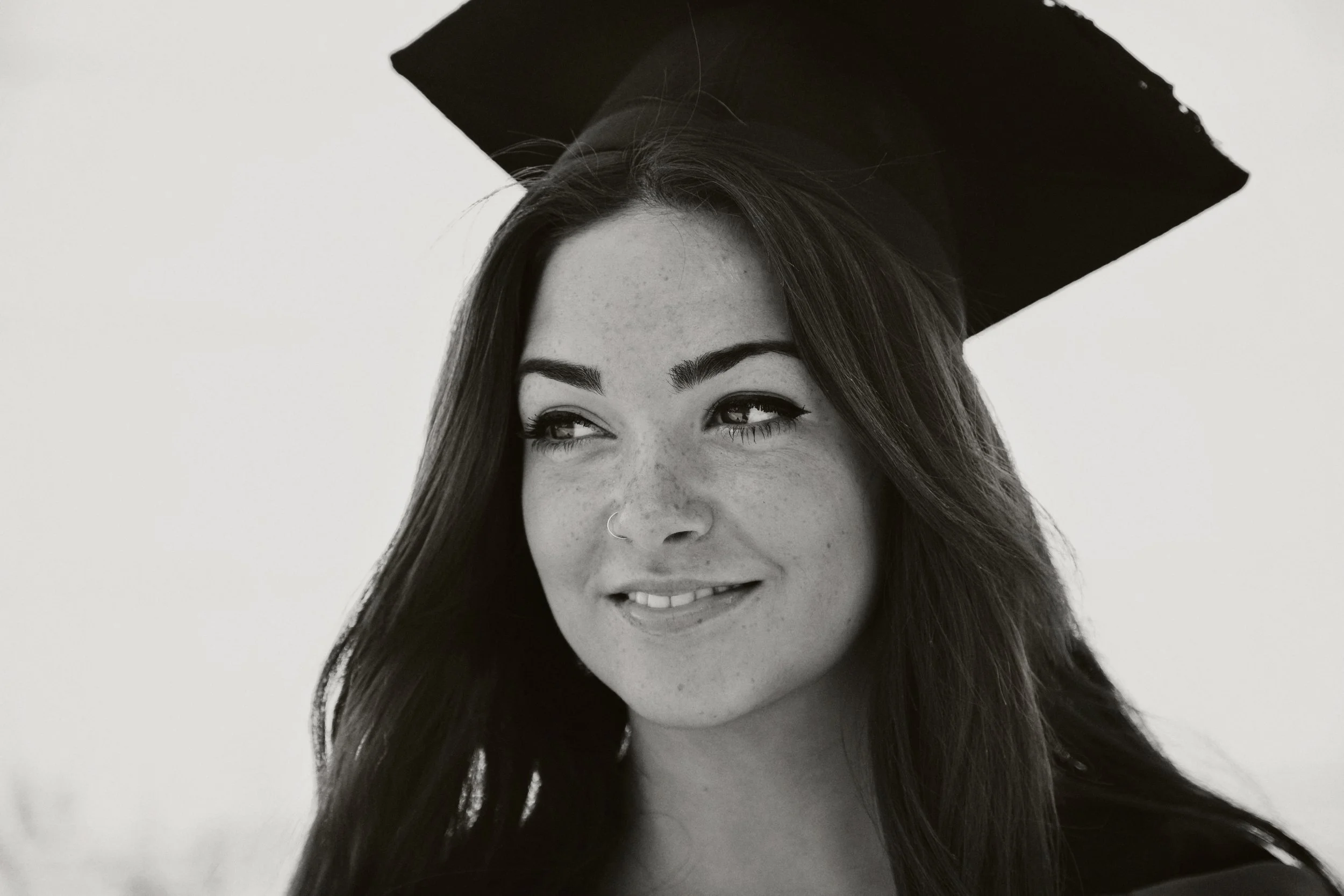 A young woman with long hair, a nose ring, and freckles, wearing a graduation cap, smiling and looking off to the side, in a black and white photo.