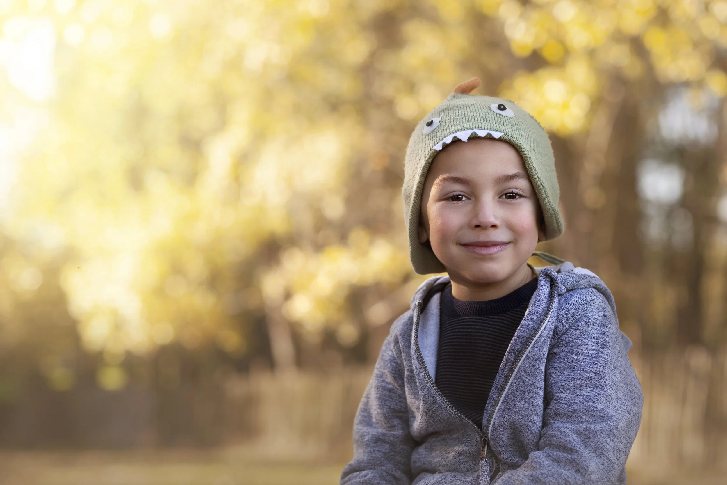 Young boy smiling sitting outside in the fall/winter with a dinosaur hat on - Luisa Young Photography 