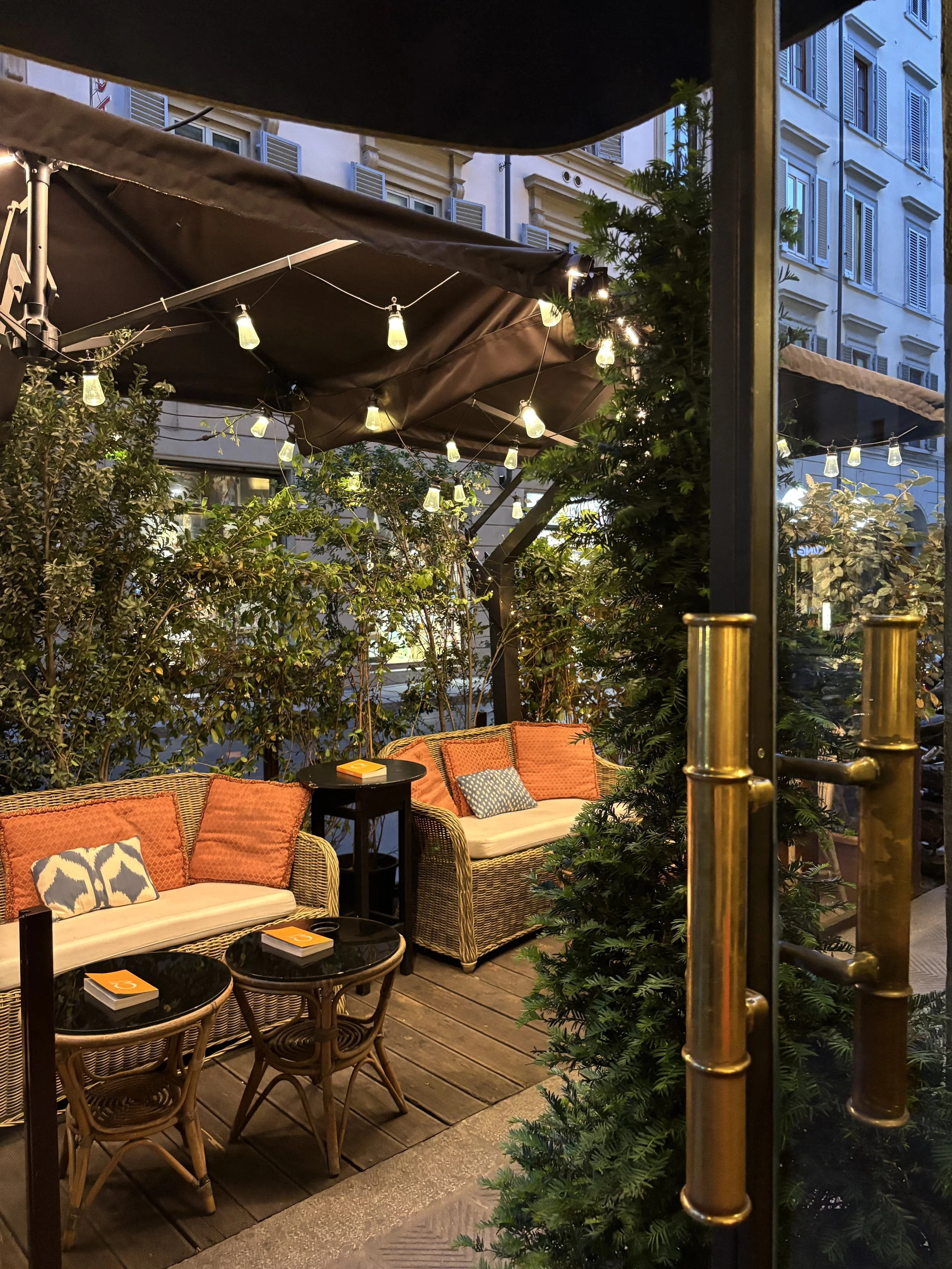 Un espace extérieur de café ou restaurant avec canapés en rotin, coussins orange et décorations lumineuses, entouré de végétation et de bâtiments urbains en arrière-plan, en soirée.