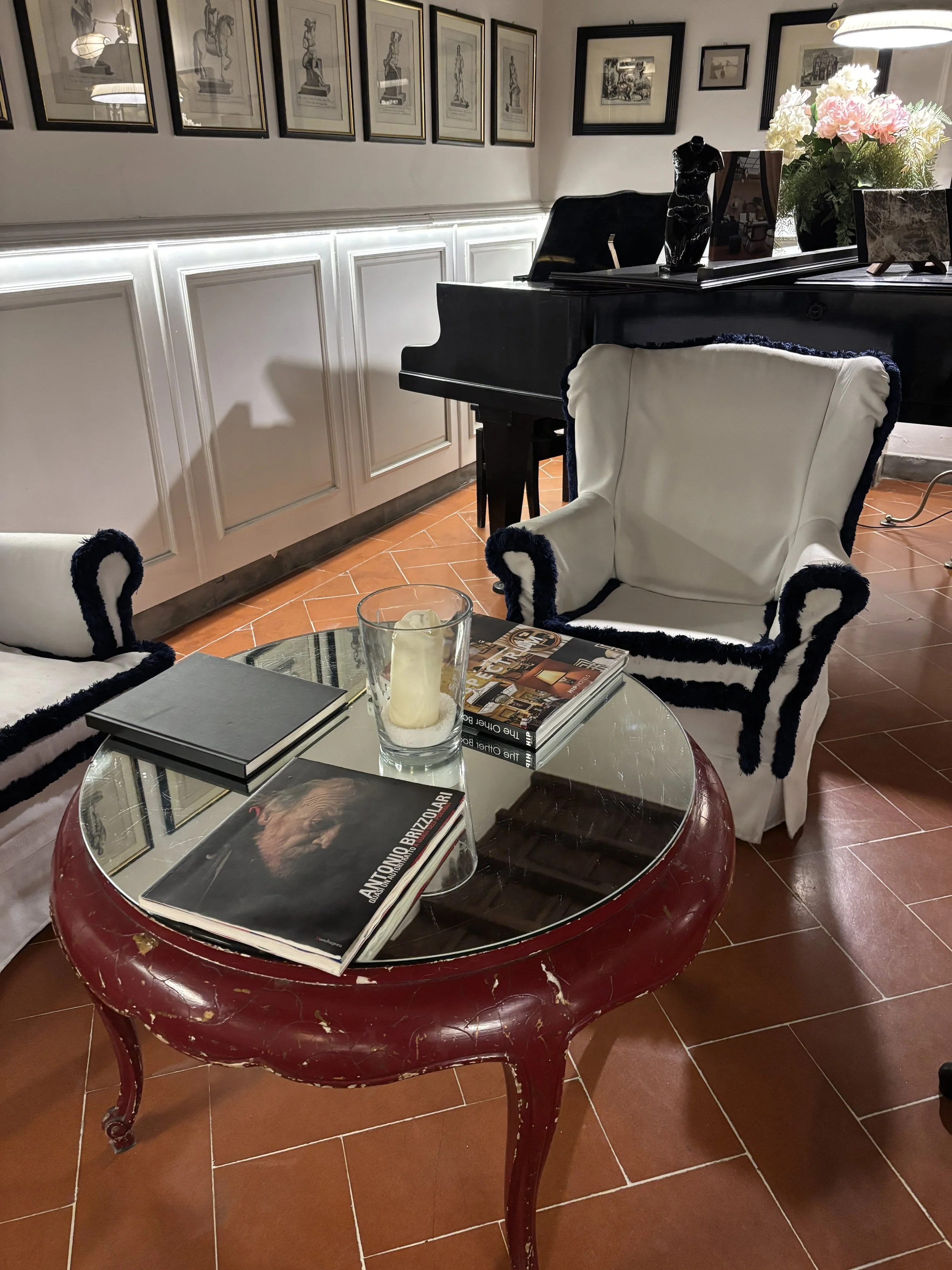 Un salon avec une table ronde en bois rouge, des magazines, une bougie dans un verre transparent, des fauteuils en tissu blanc avec des contours en velours noir. En arrière-plan, un piano noir avec des cadres contenant des dessins ou des photographies accrochés au mur, des fleurs blanches et roses dans un vase, et des décorations artistiques.