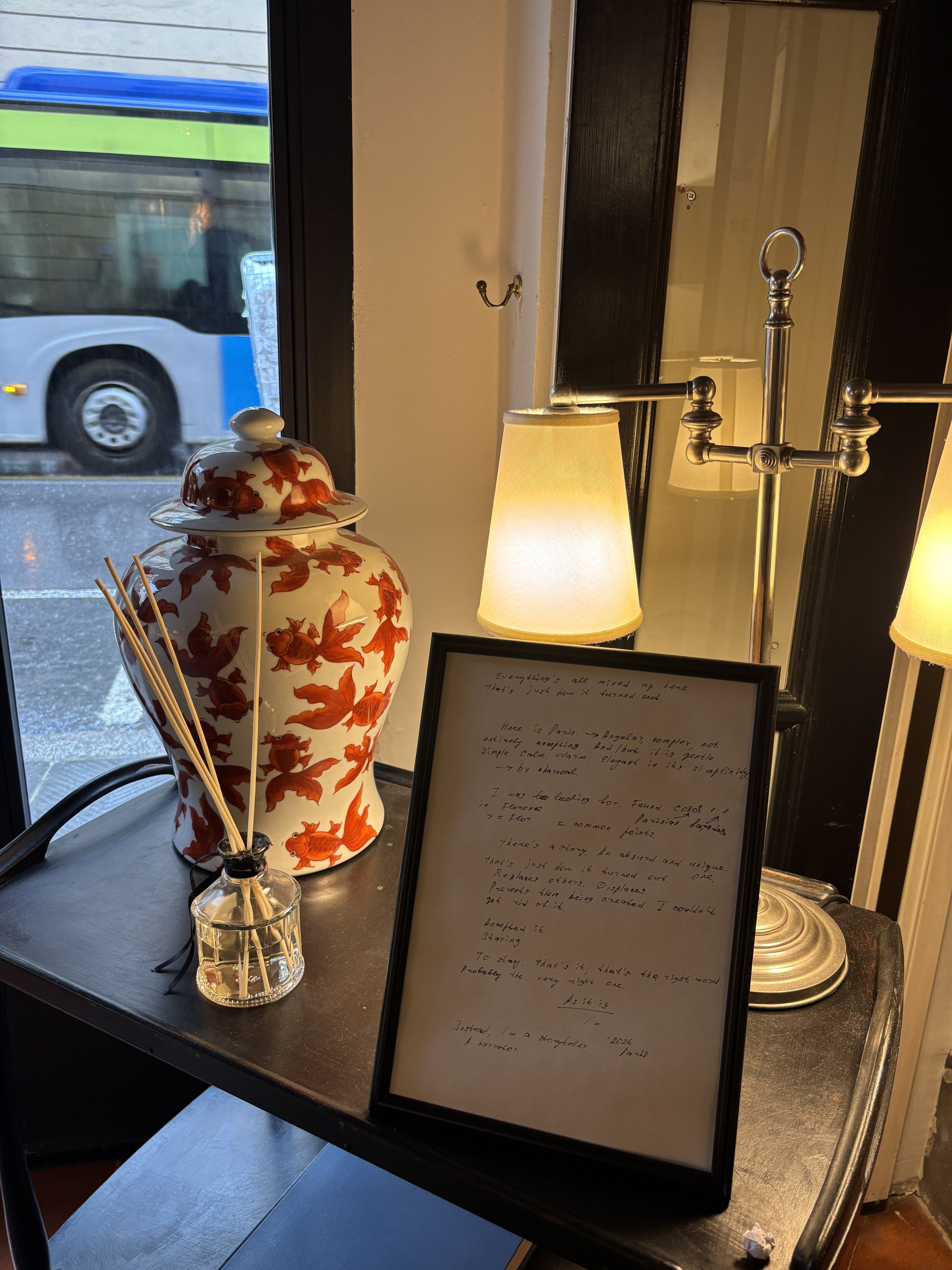 Une lampe en porcelaine blanche avec des poissons orange, un cadre avec un texte manuscrit en anglais, une lampe à abat-jour beige, et un vase avec des brochettes en bois, sur une table près d'une fenêtre.
