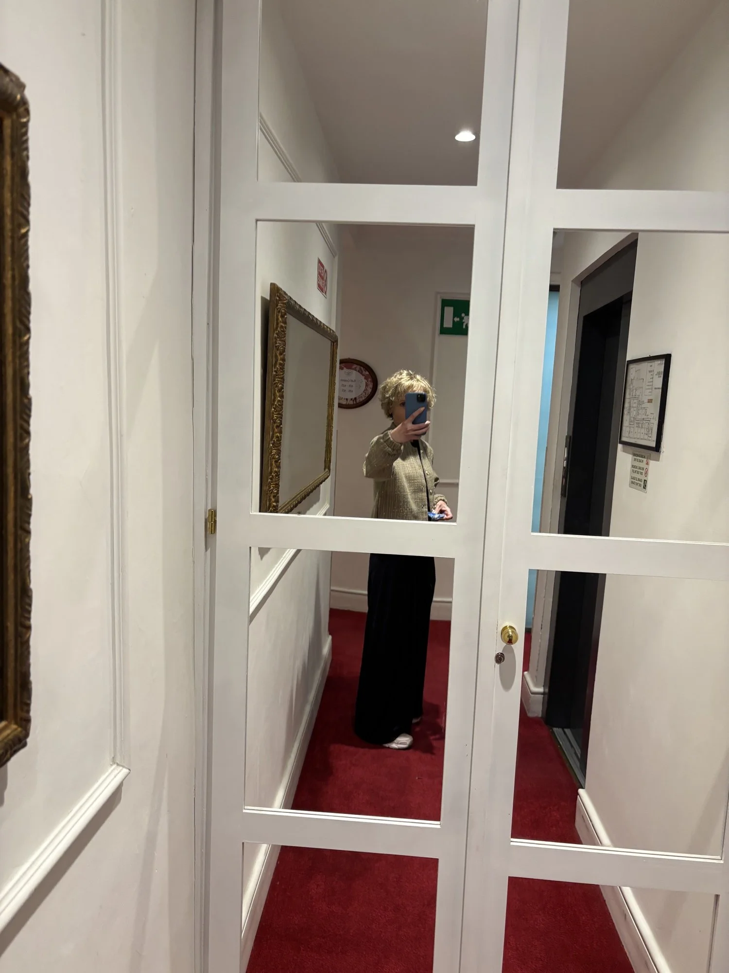 Une femme avec des cheveux blonds et frisés, portant un pull beige et un pantalon noir, se tient dans un couloir d'hôtel ou de bâtiment, prenant une photo d'elle-même avec un téléphone portable, visible dans un miroir.
