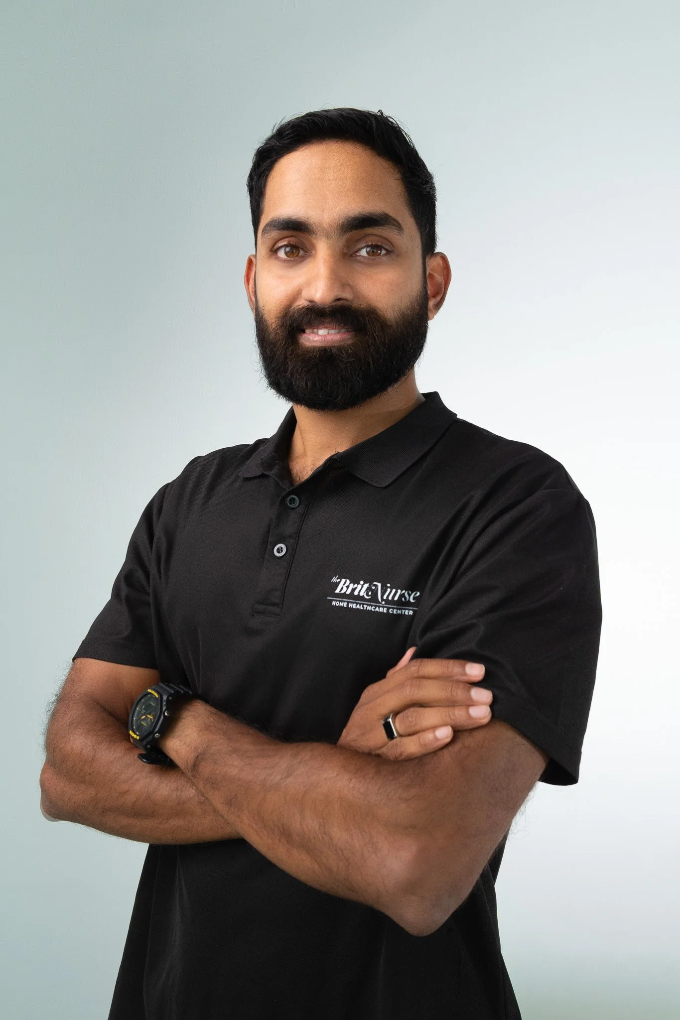 A man with short dark hair and a beard, wearing a black polo shirt with a logo and text, standing with arms crossed against a light-colored background.