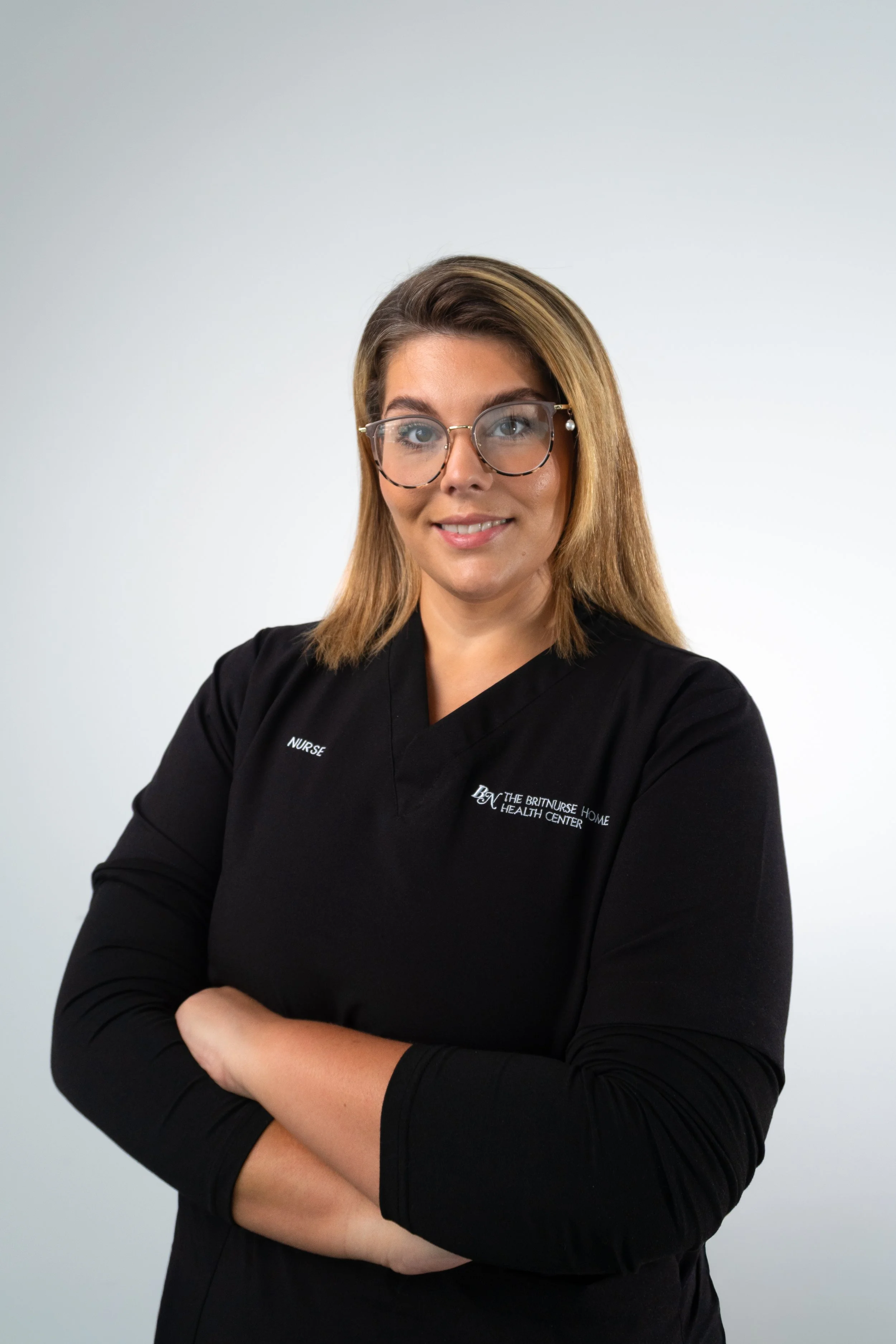 A female nurse with shoulder-length blonde hair, wearing glasses, a black uniform with company logo, standing with arms crossed against a neutral background.