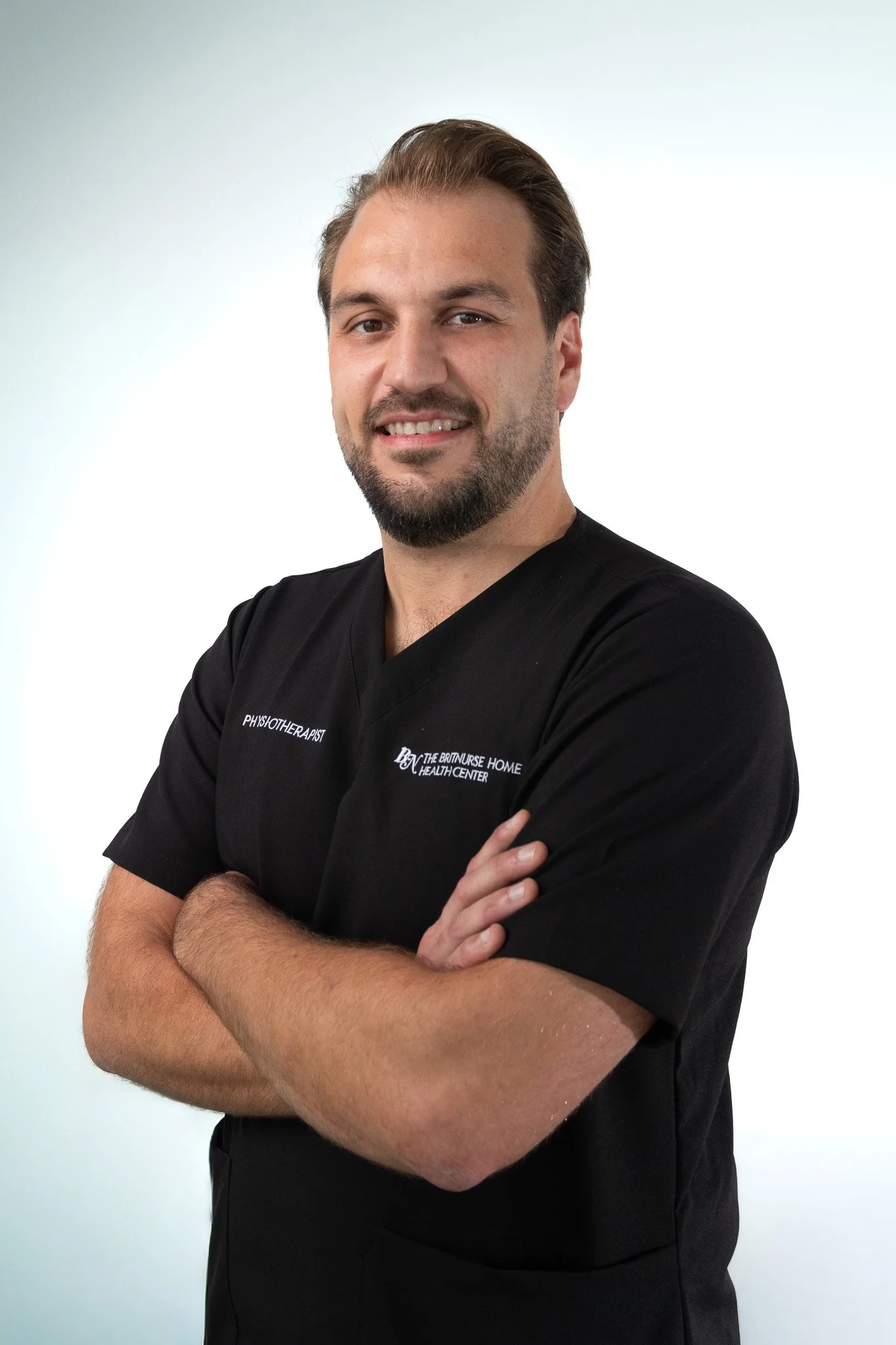 A man with a beard and short hair, smiling and crossing his arms, wearing a black shirt with 'Physiotherapist' embroidered on it, standing against a plain background.