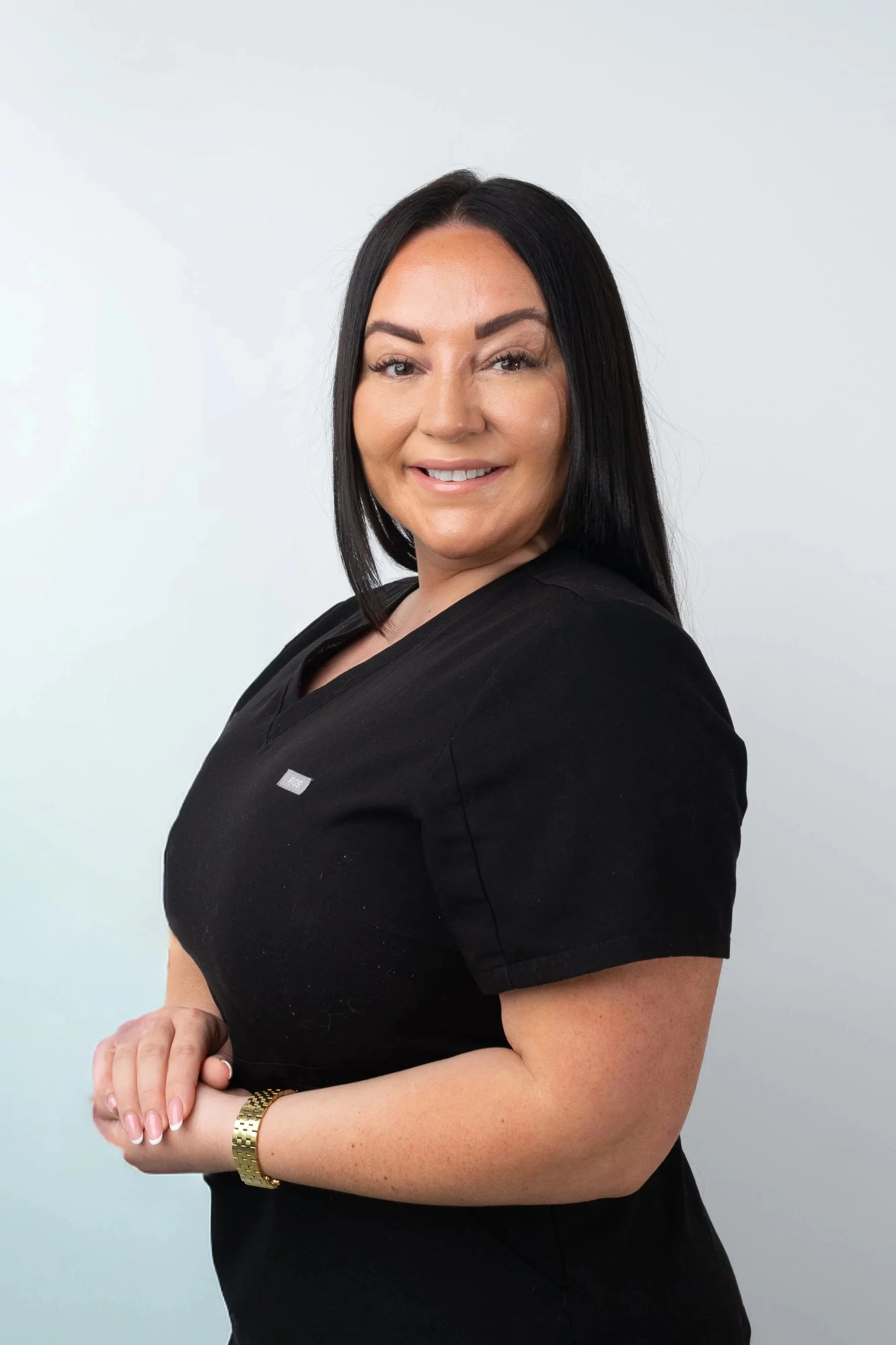 A woman with long black hair smiling, wearing a black shirt and a gold watch, standing against a plain light background.